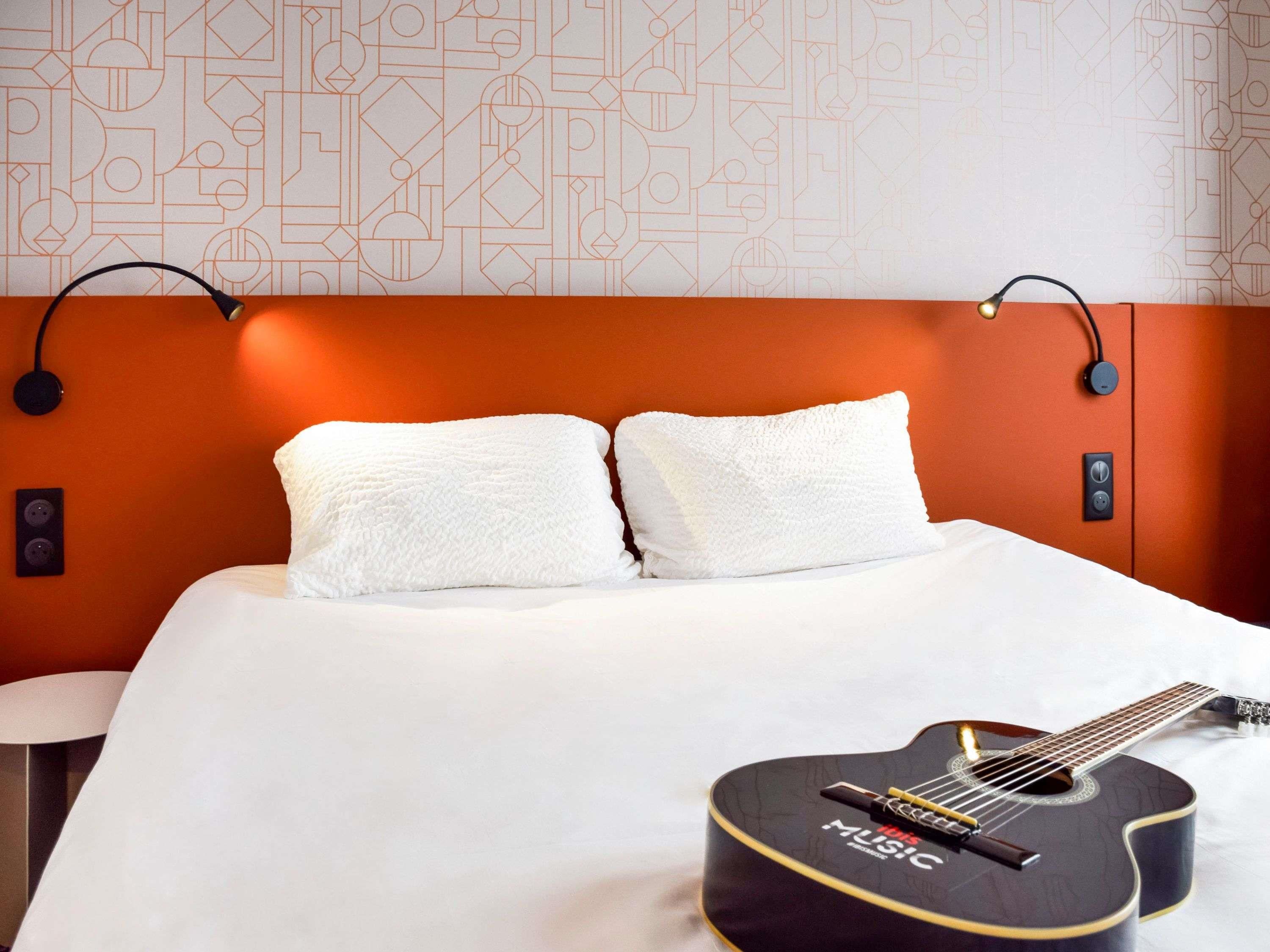 A hotel room with a bed, two pillows, a guitar, and a wall with geometric patterns.