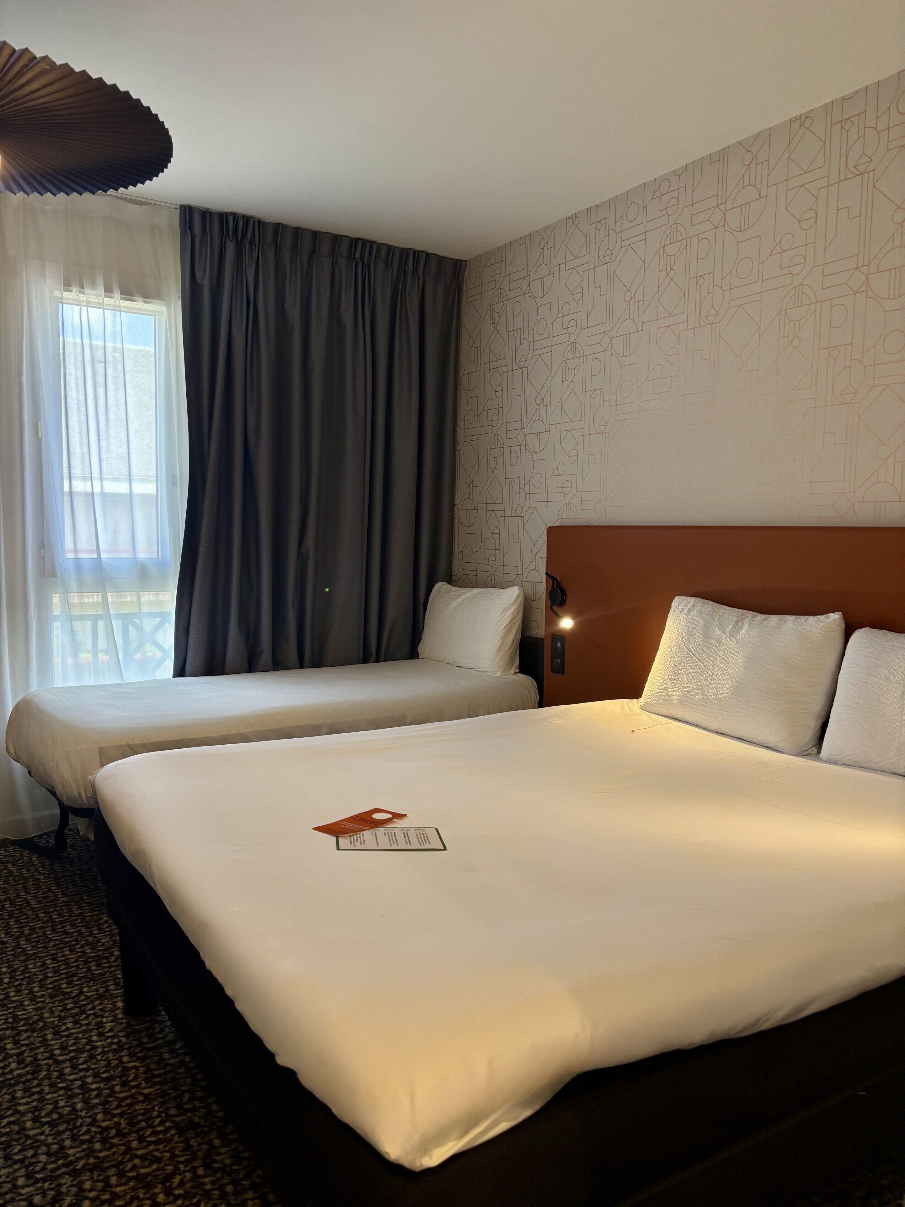 A hotel room with two beds, a large window with curtains, a patterned wall, and a headboard with a light fixture.