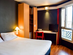 A hotel room with a bed, a desk with a TV, a chair, a lamp, and a window with a view. - ibis Rodez Centre Cathédrale (Rodez)