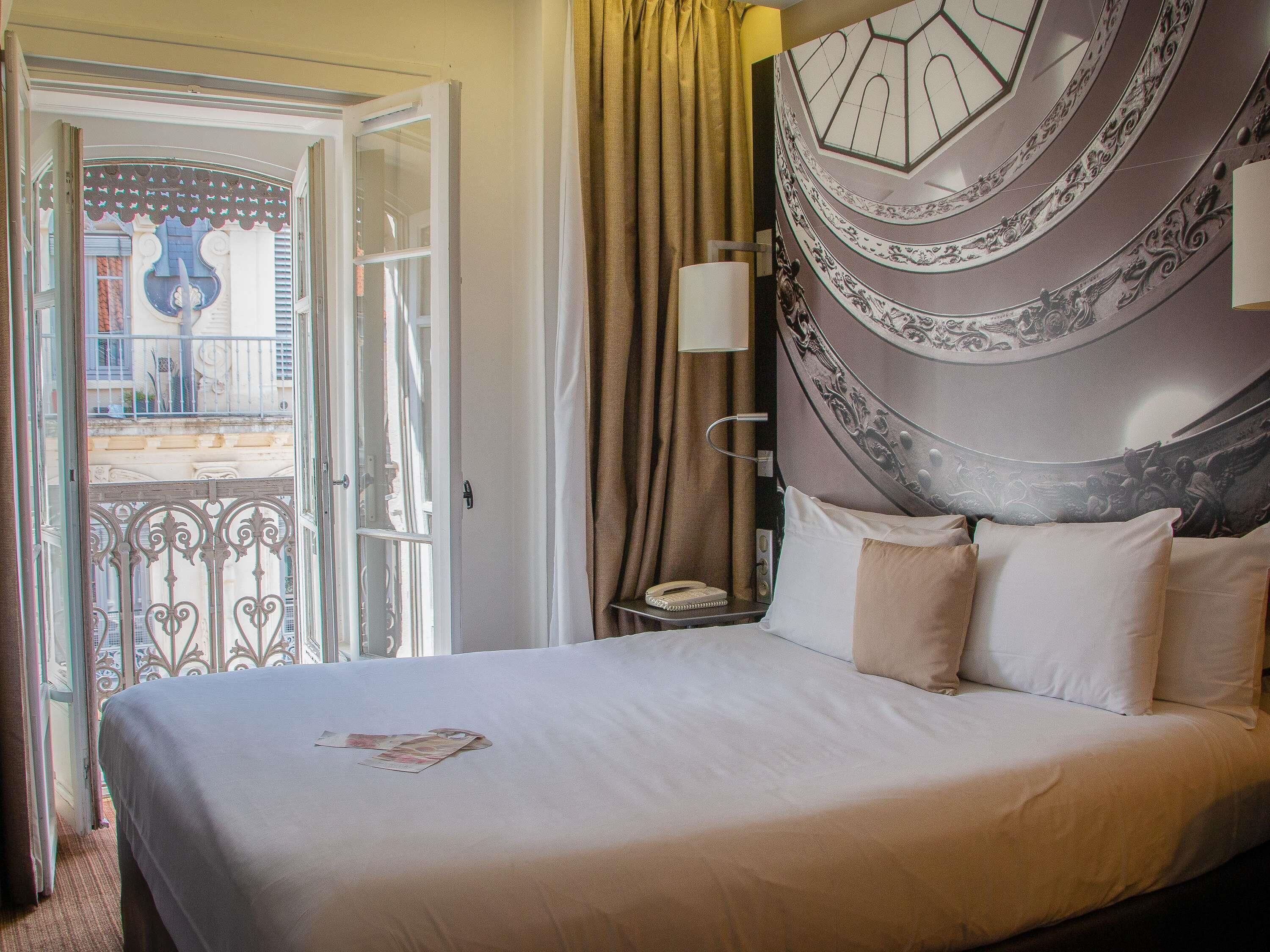 A hotel room with a bed, a nightstand, a lamp, and a view of the outside.