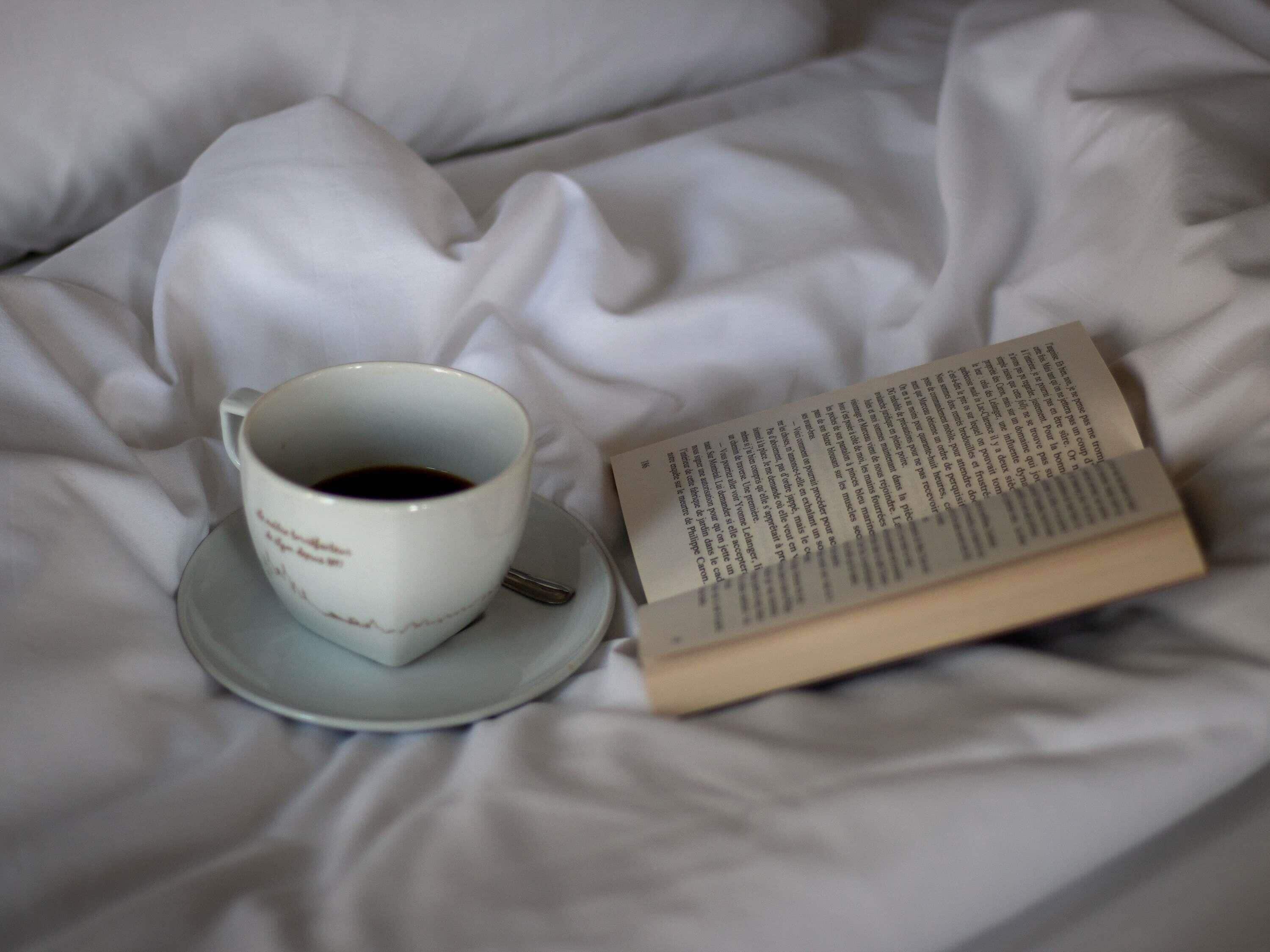 Eine Tasse Kaffee auf einem Untertasse und ein offenes Buch auf einem Bett.