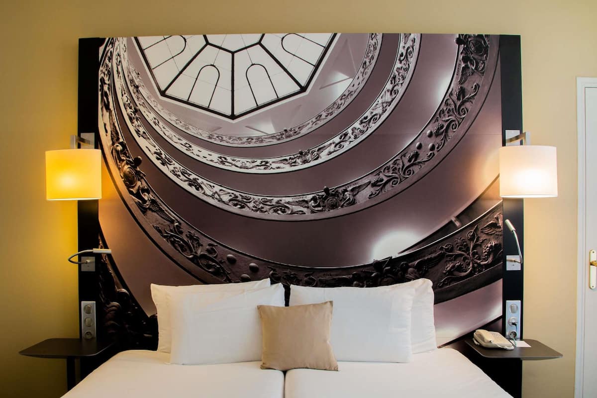 A hotel room with a large bed, two bedside lamps, a wall-mounted telephone, and a decorative ceiling.