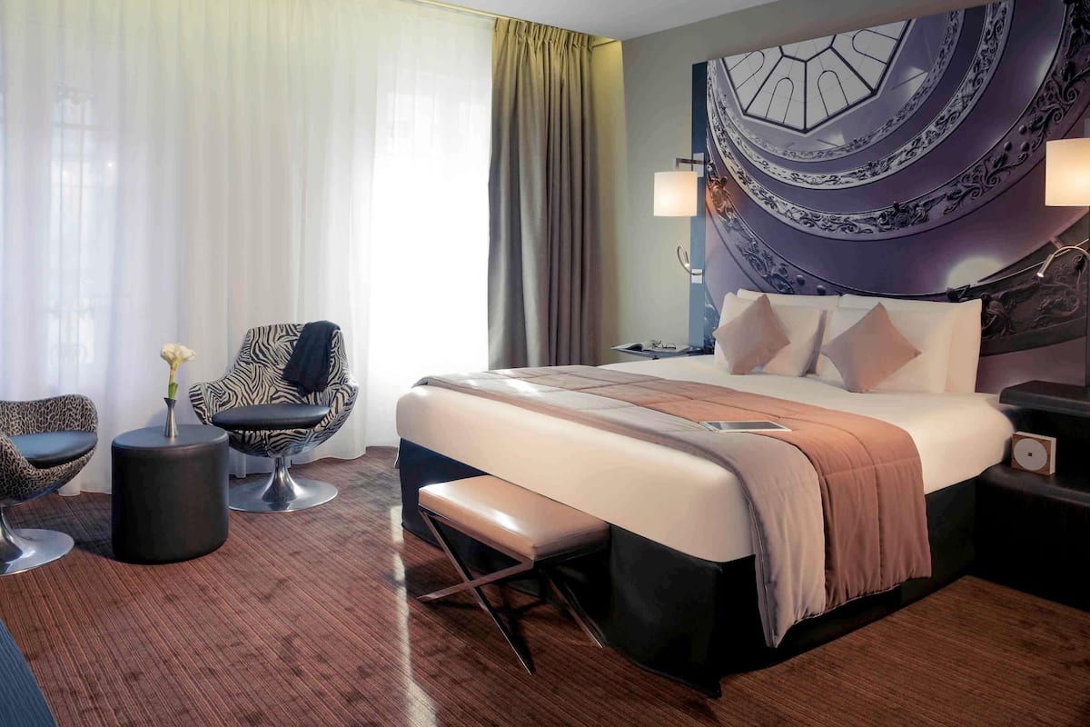 A modern hotel room with a large bed, a seating area with a chair and a small table, a nightstand, and a decorative wall piece.