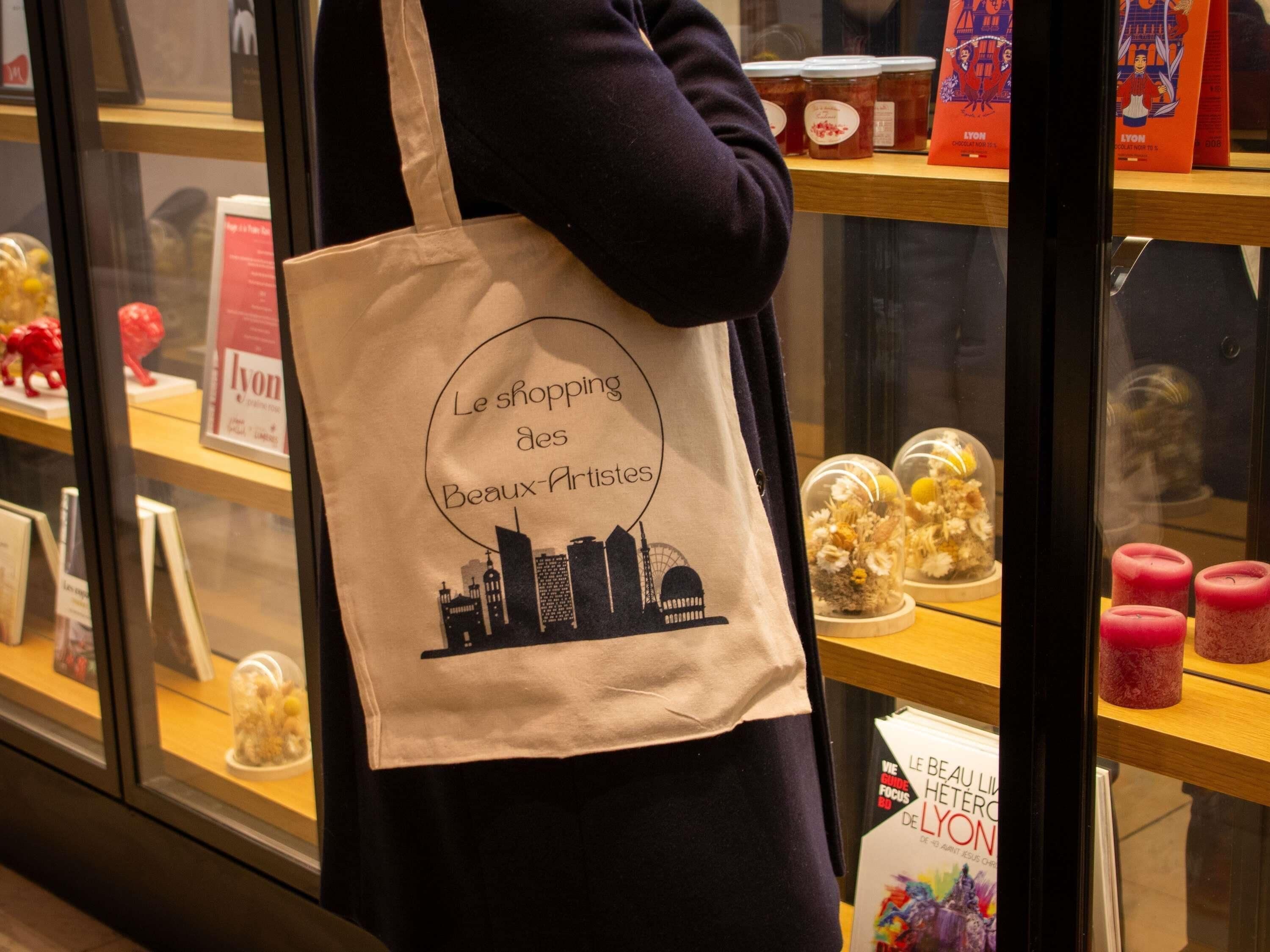 Eine Person hält eine Stofftasche mit dem Aufdruck 'Le shopping des Beaux-Arts' in der Hand und steht vor einer Vitrine mit verschiedenen Gegenständen.