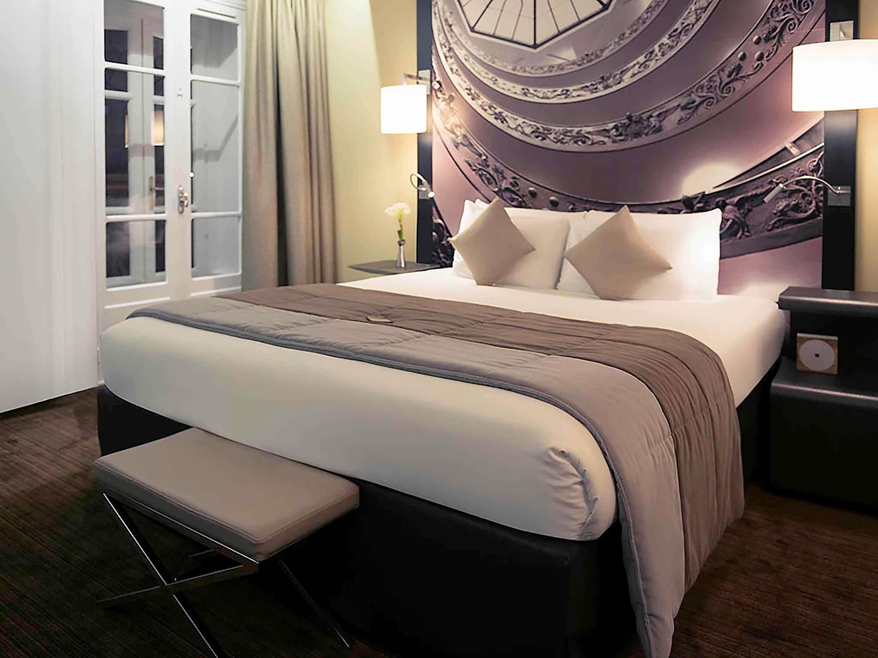 A hotel room with a large bed, a bedside table, a lamp, and a window with curtains.