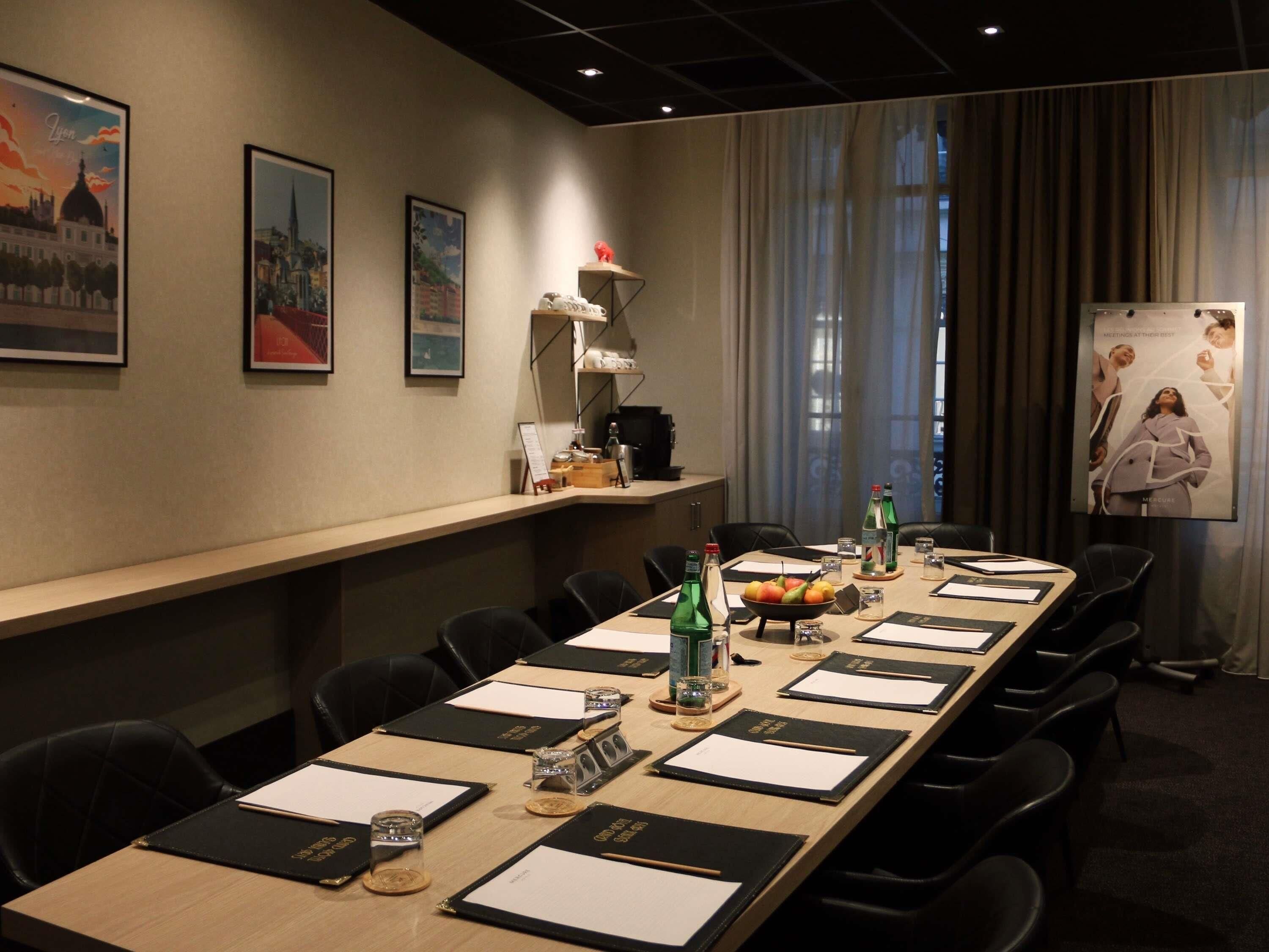 A conference room with a long table, black chairs, framed pictures on the wall, a projector screen, and a window with curtains.