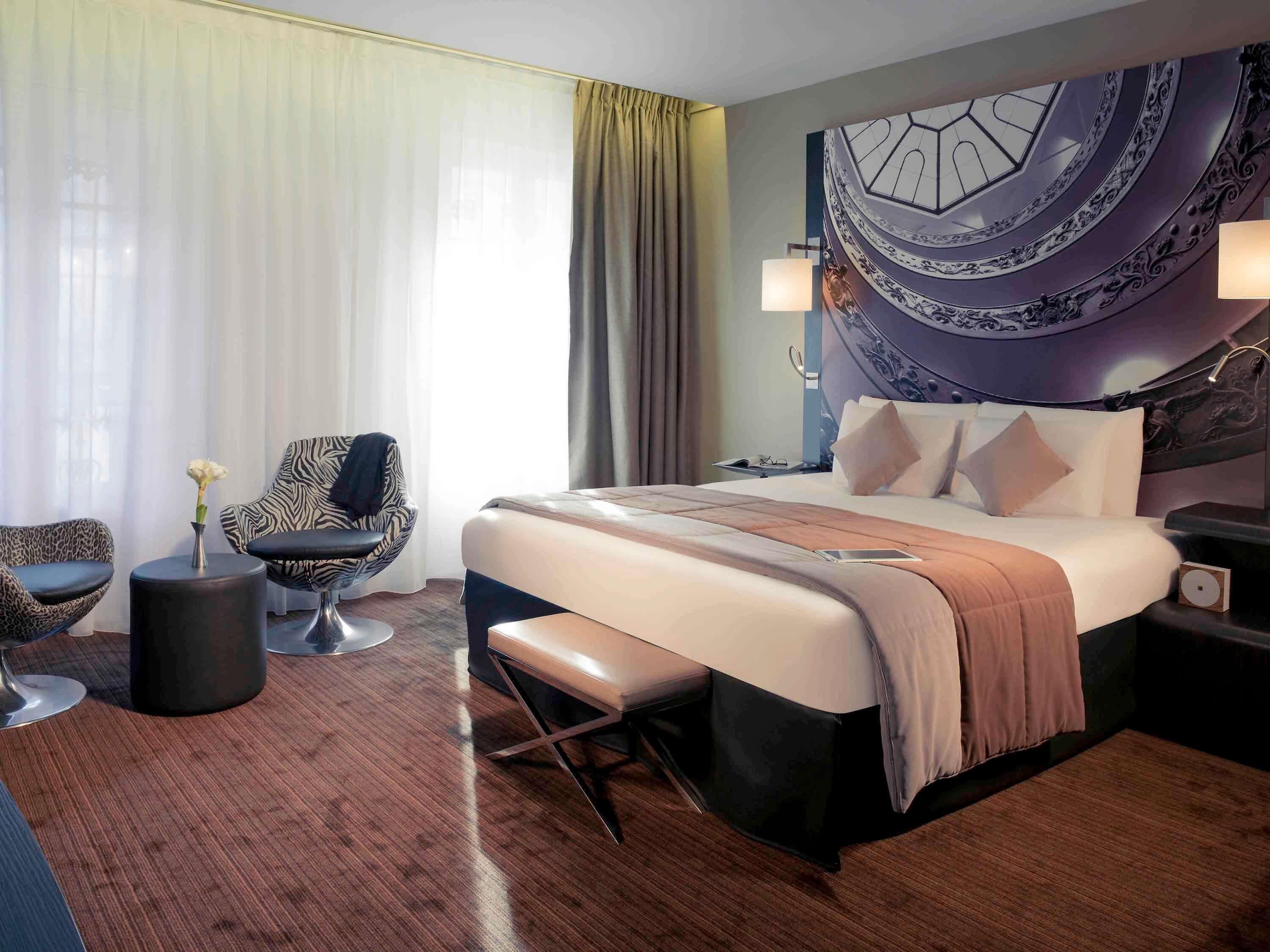 A modern hotel room with a large bed, a seating area with a chair and a small table, a nightstand, and a decorative wall piece.