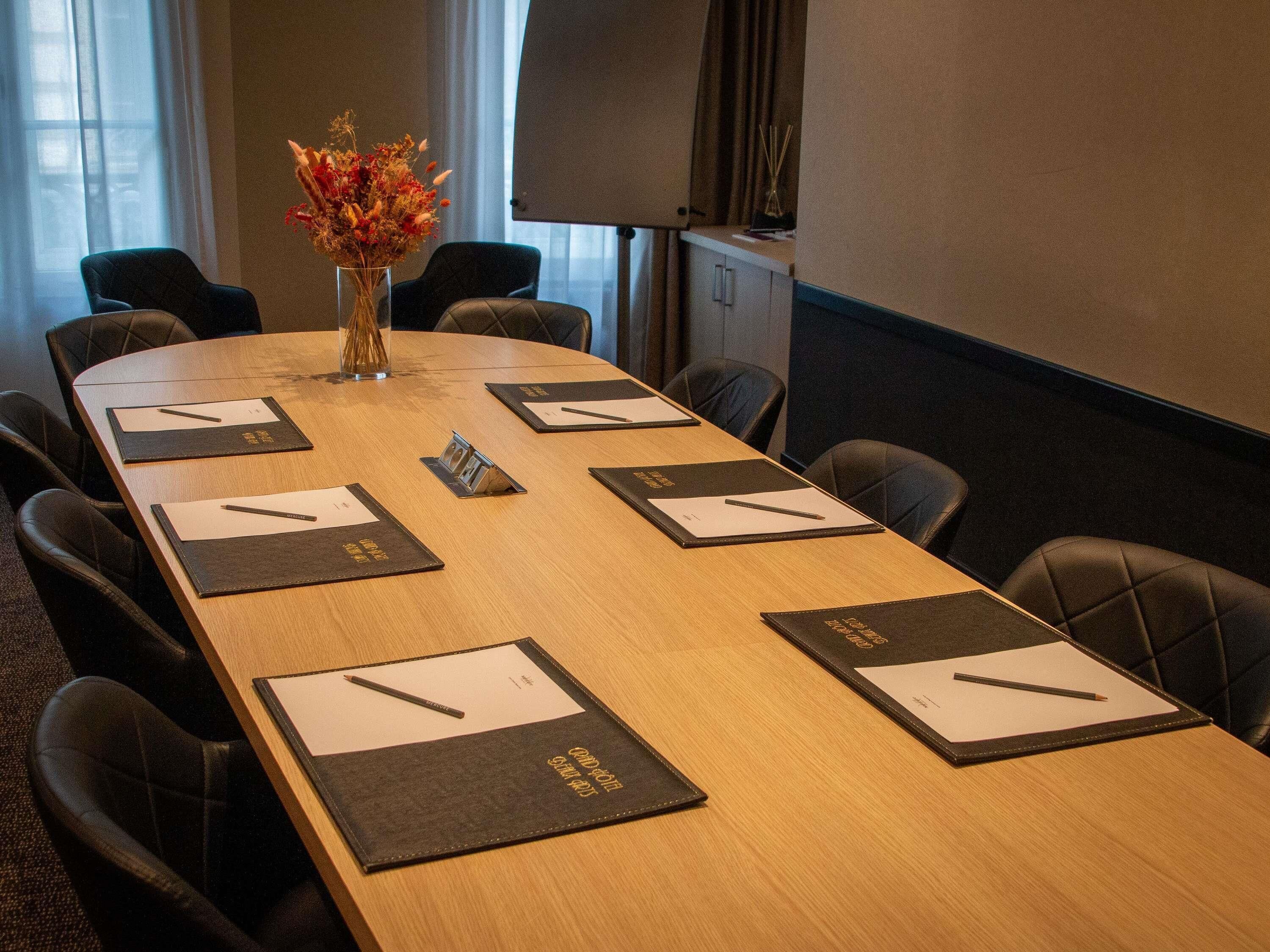 A conference room with a long table, black leather chairs, a projector screen, and a vase with flowers.