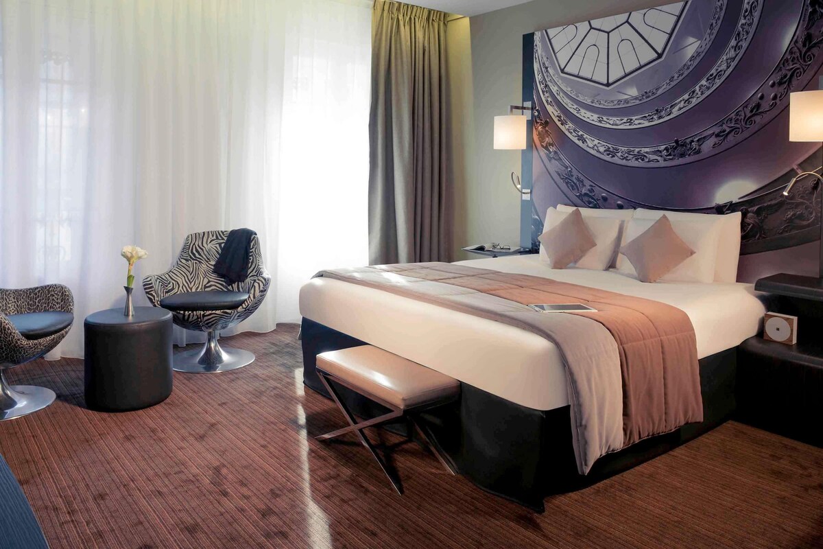 A modern hotel room with a large bed, a seating area with a chair and a small table, a nightstand, and a decorative wall piece.