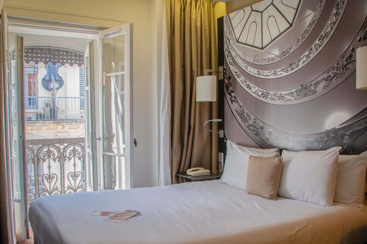 A hotel room with a bed, a nightstand, a lamp, and a view of the outside.