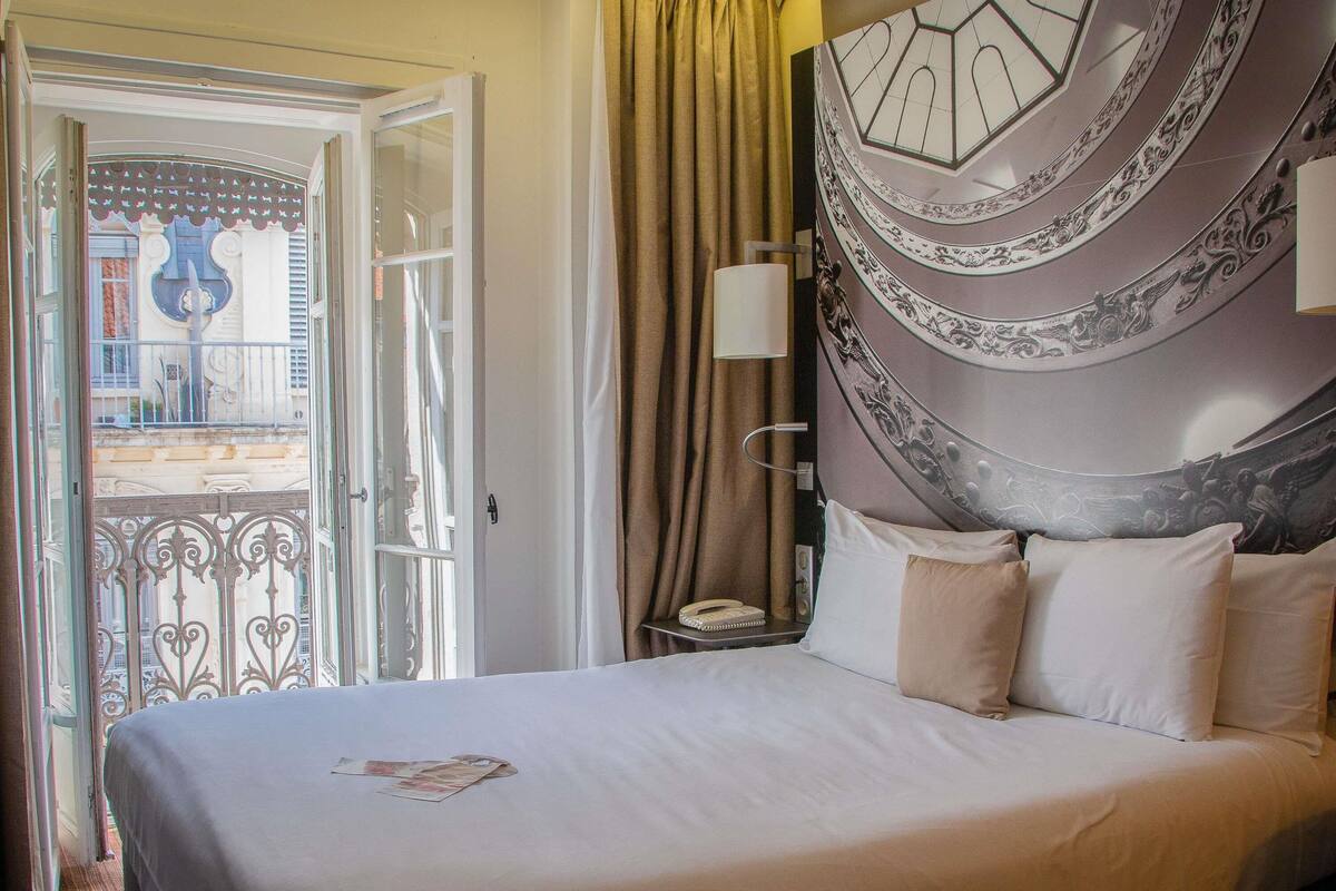 A hotel room with a bed, a nightstand, a lamp, and a view of the outside.