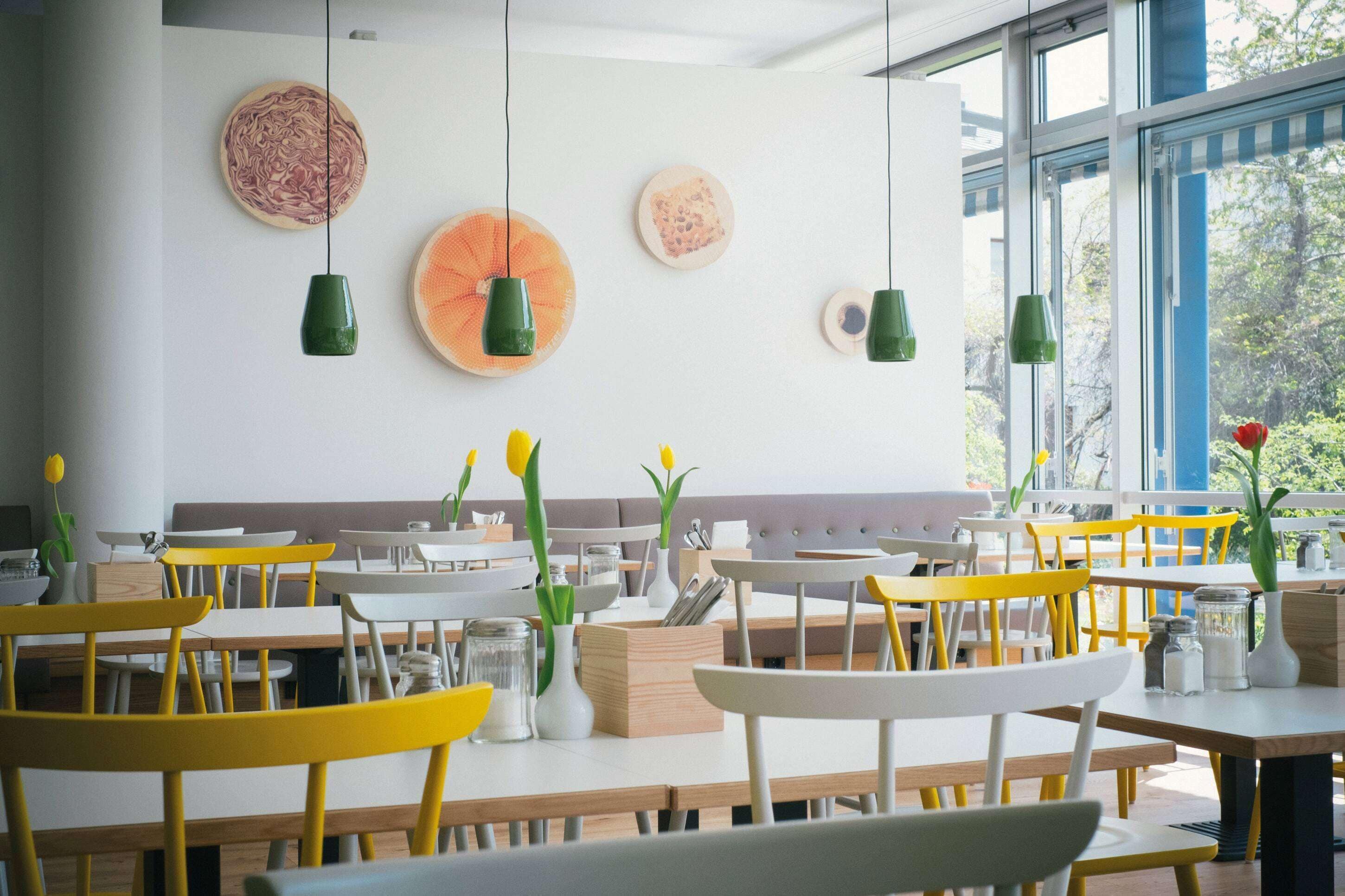 A modern dining area with wooden tables and yellow chairs, a grey cushioned bench, and green pendant lights.