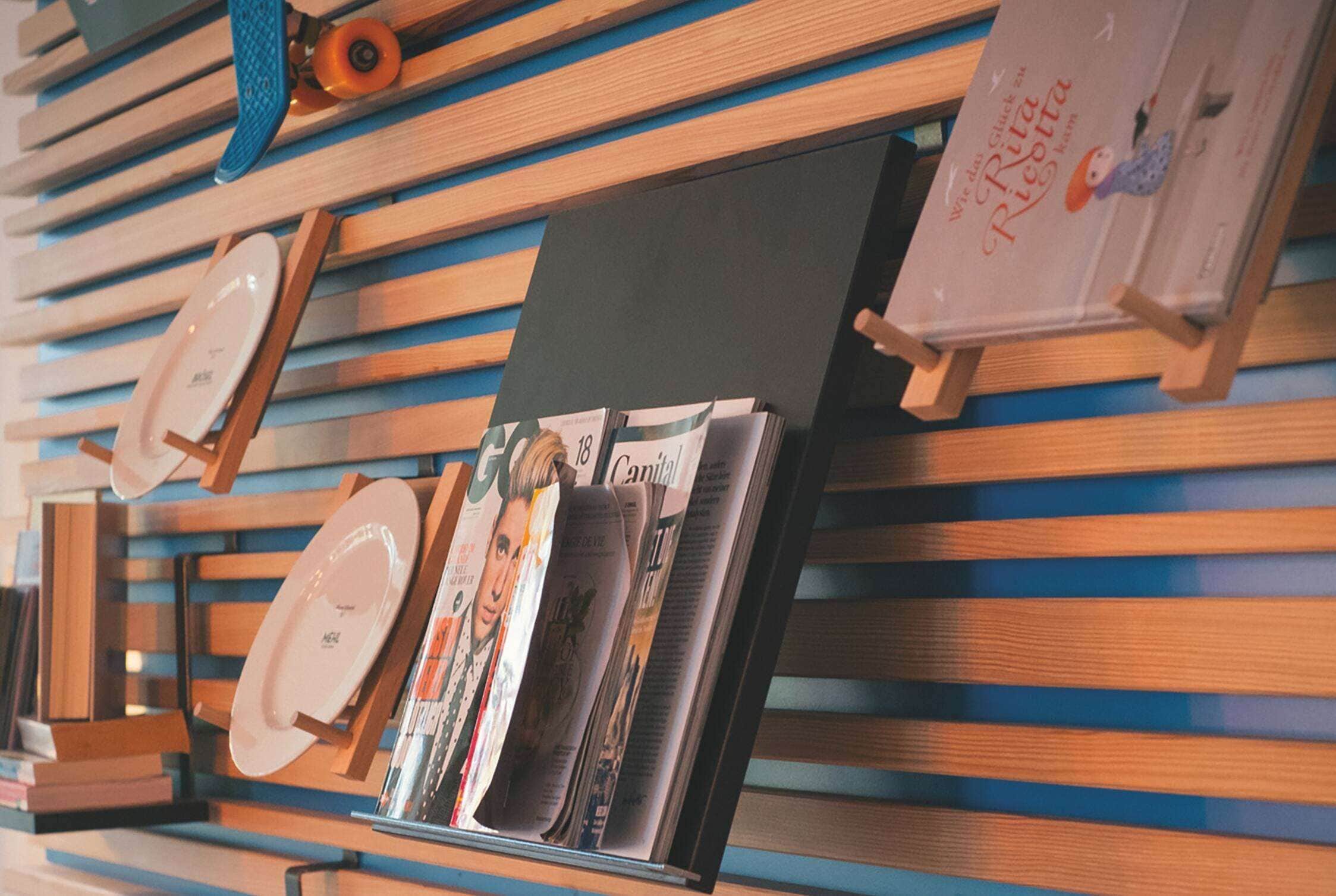 A magazine rack with various publications, a plate, and a book titled "Rita Ricotta" on a wooden slatted wall.