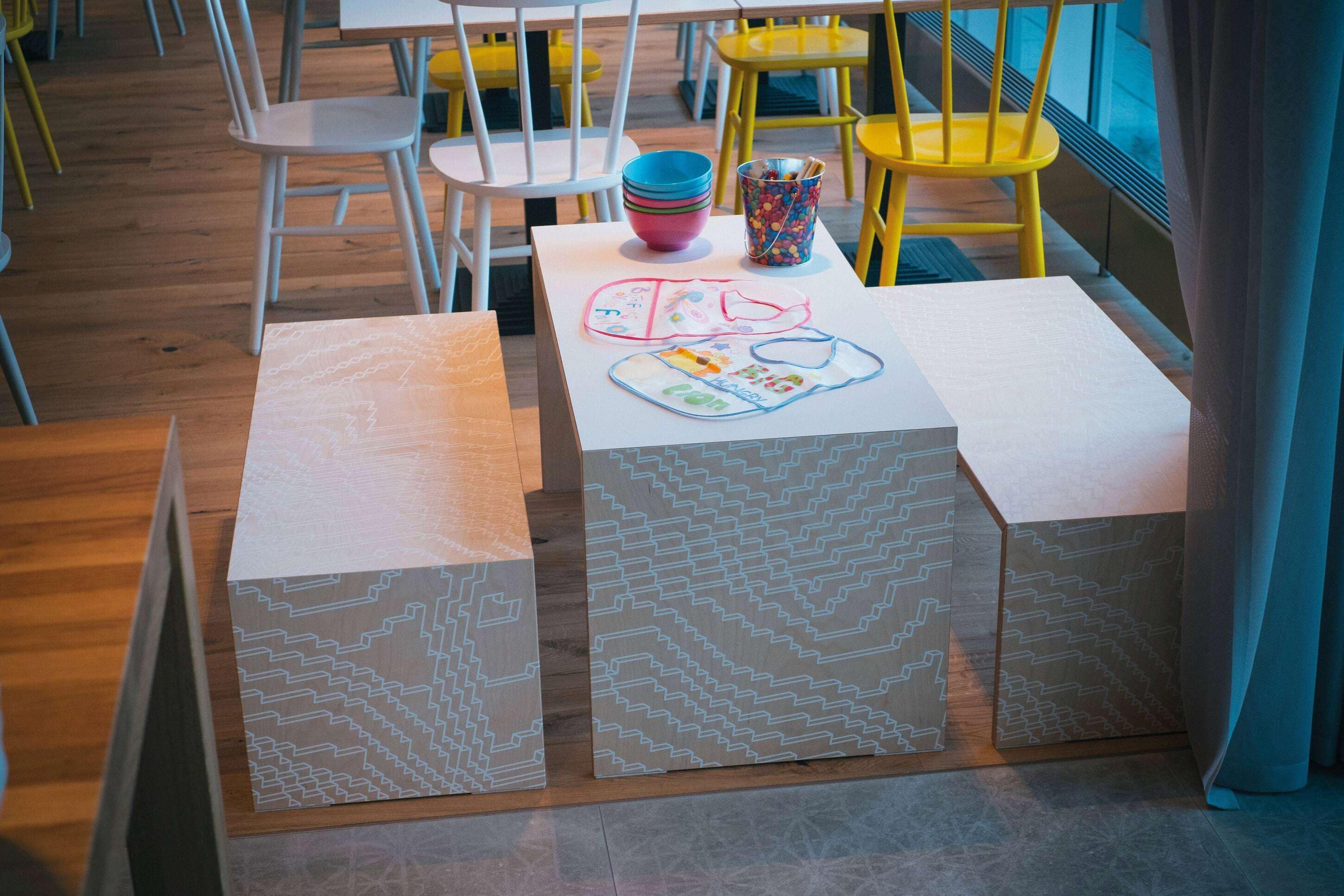 A dining area with tables and chairs, some with colorful cushions. There are patterned boxes on the tables.