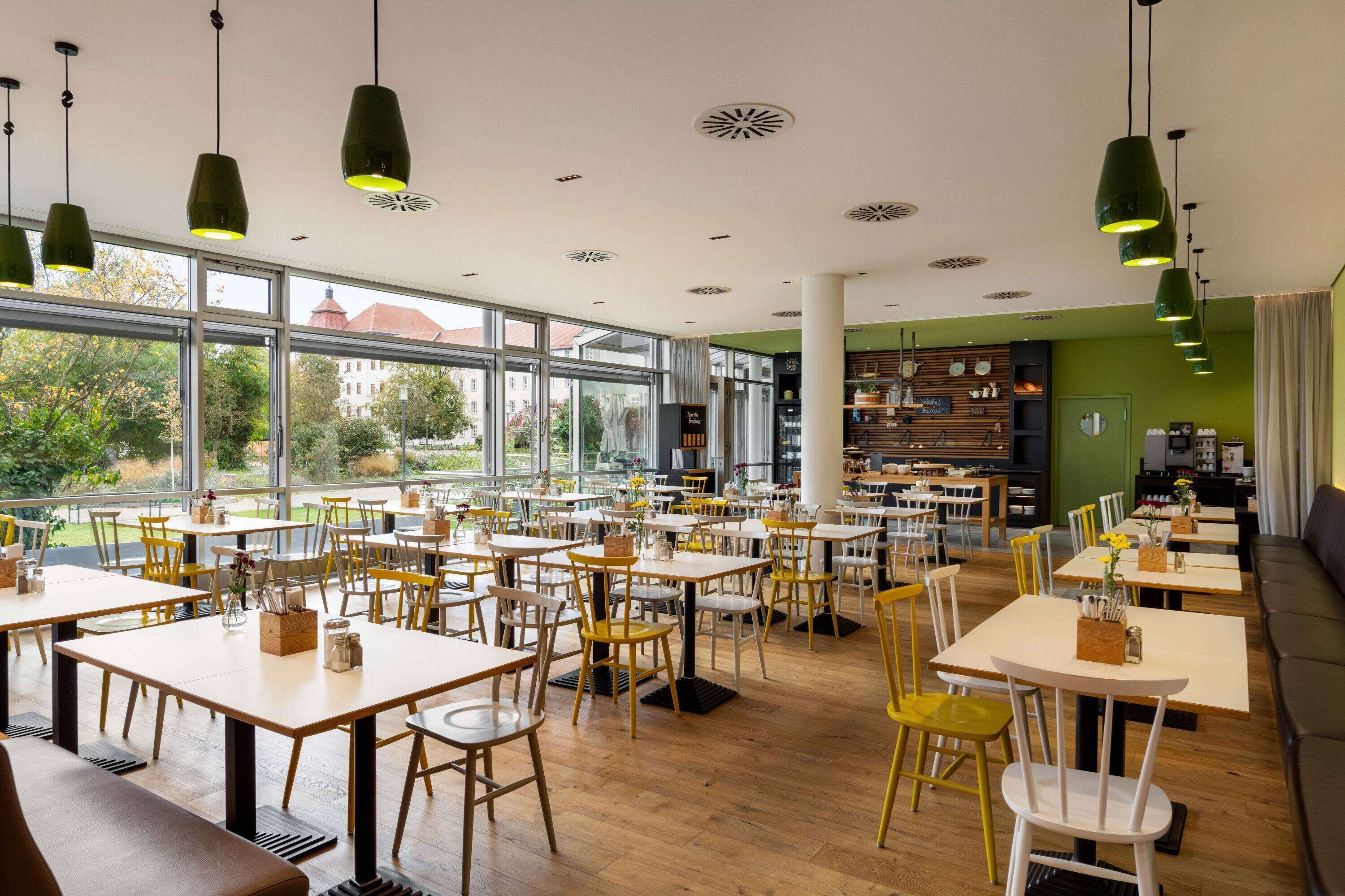 Un comedor amplio con varias mesas y sillas, ventanas grandes que ofrecen vistas al exterior y una pared de acento verde.