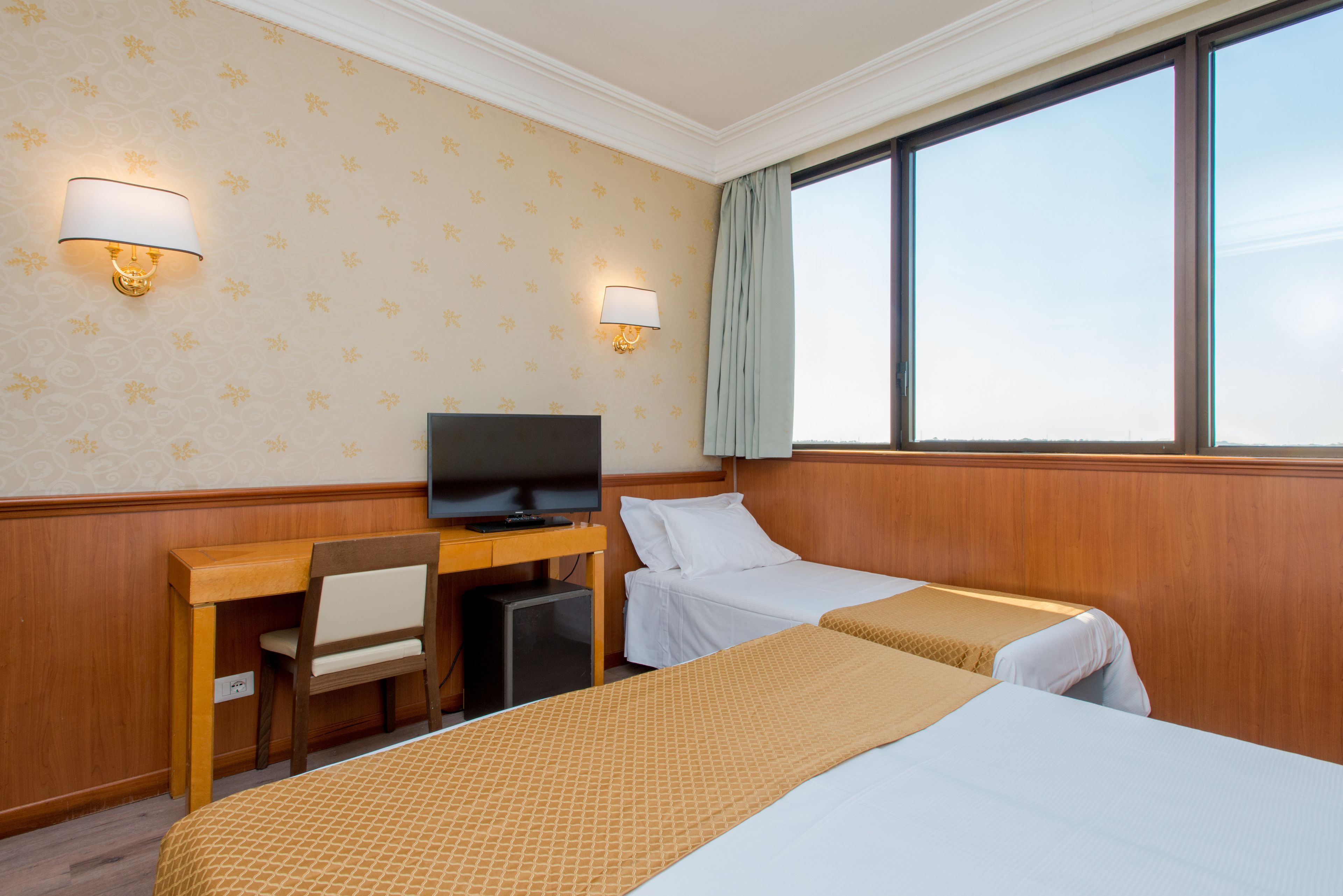 A hotel room with two beds, a desk with a television, and two wall-mounted lamps.