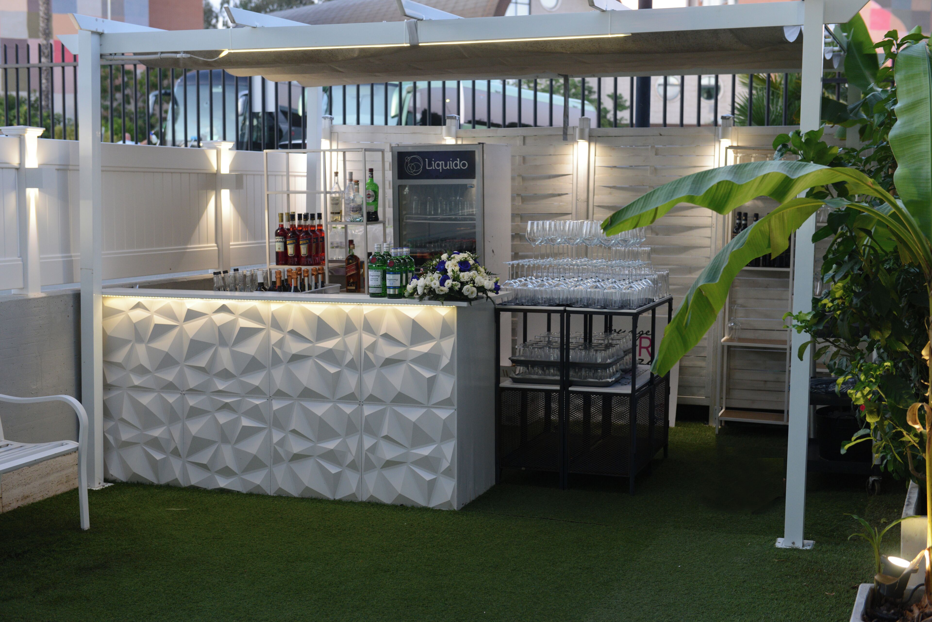 A bar area with a white geometric wall, a refrigerated display, and a variety of bottles and glasses.