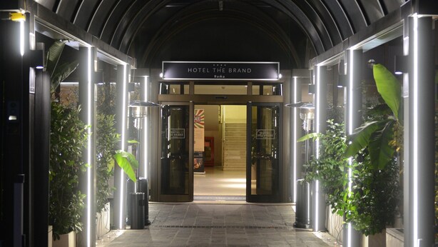 Entrada de hotel con diseño moderno en arco, iluminada por luces, y un cartel que dice 'HOTEL THE BRAND'.