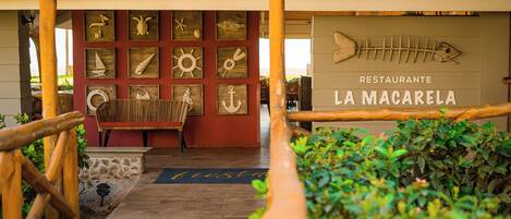 A wooden pathway leading to a restaurant named 'LA MACARELA' with a thatched roof and nautical-themed decorations.
