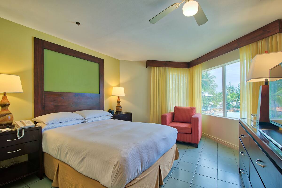 A hotel room with a large bed, two bedside lamps, a wooden headboard, a red chair, a dresser, and a large window with curtains.