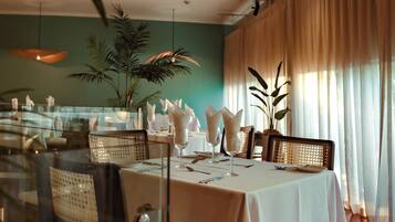 A dining area with a table set for four, surrounded by chairs, with a large plant in the foreground and a view of a window with curtains.