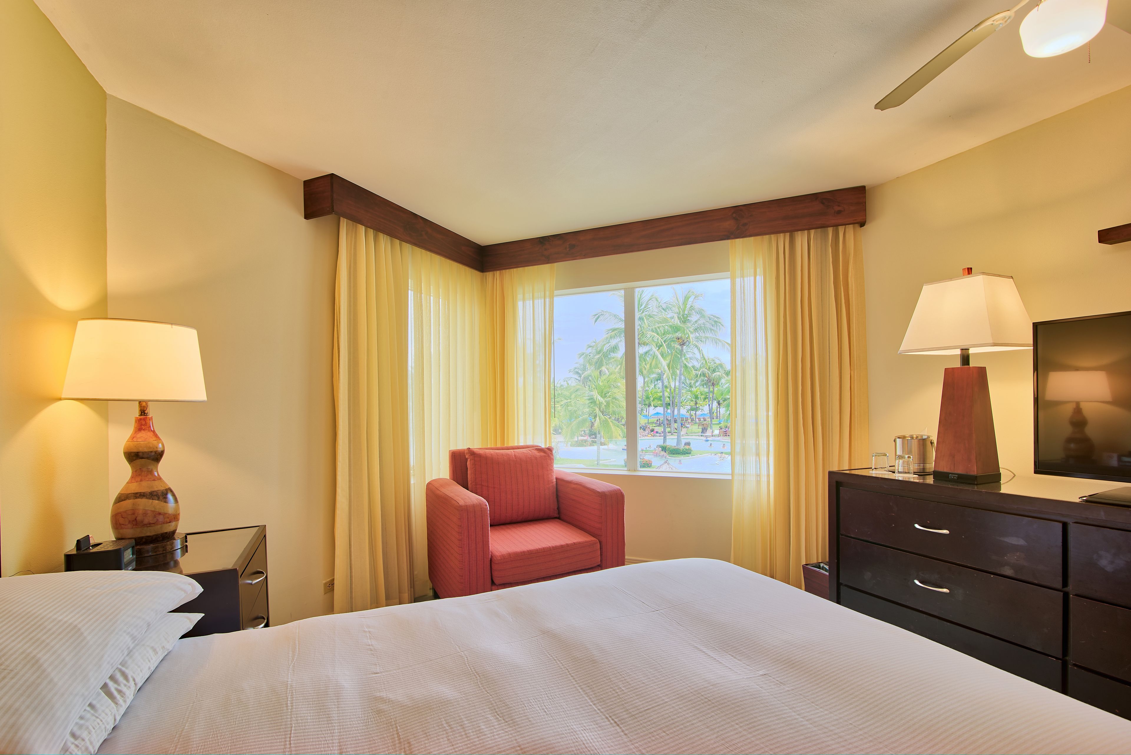 A hotel room with a bed, a red armchair, a wooden dresser, and a window with curtains.