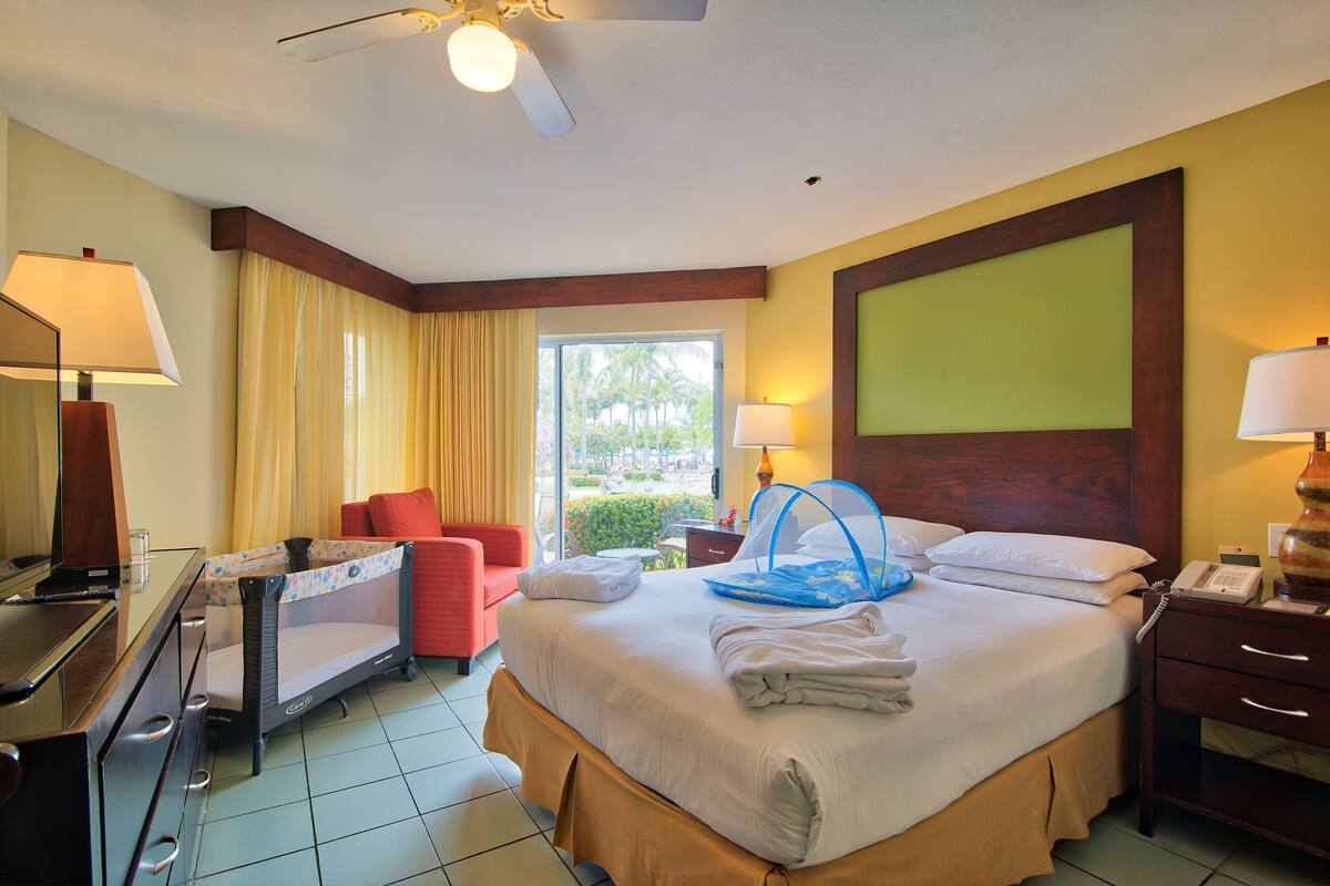 A hotel room with a bed, a desk, a chair, a desk lamp, a bedside table with a lamp, and a window with curtains.