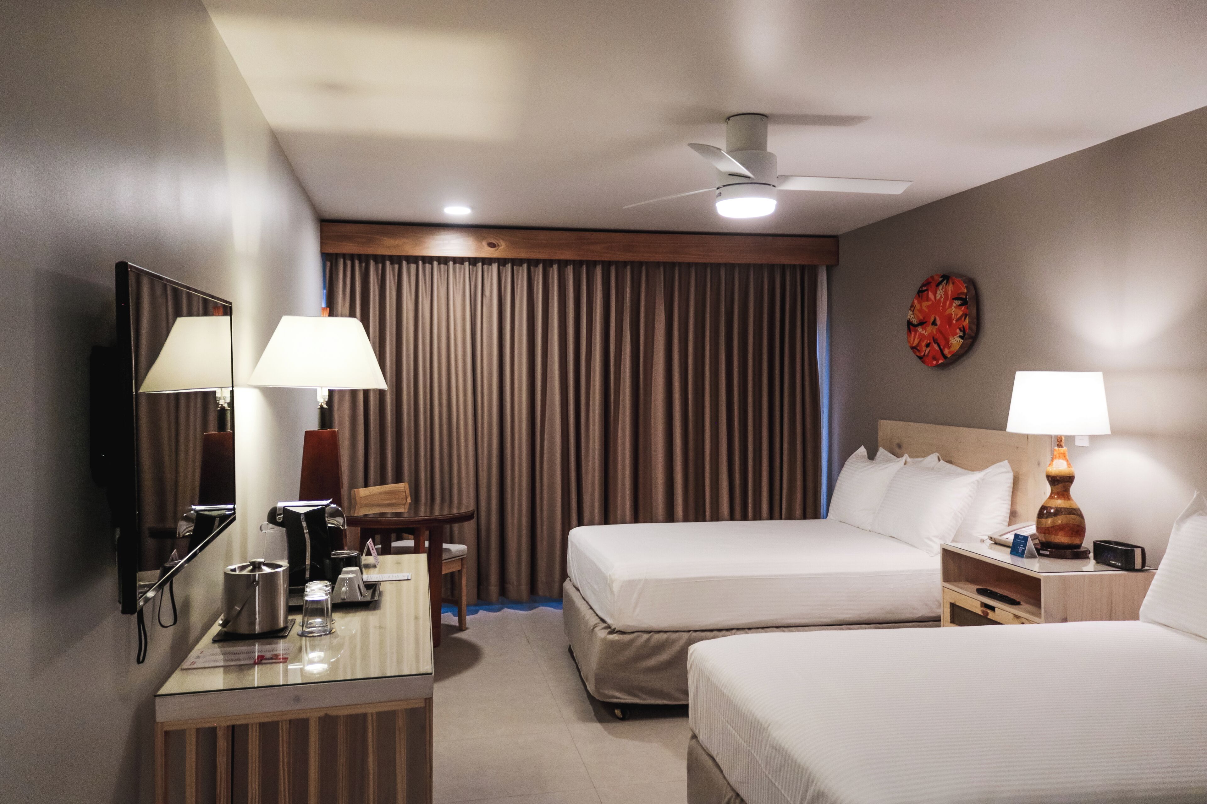 A hotel room with two beds, a desk with a lamp, a coffee maker, and a ceiling fan.