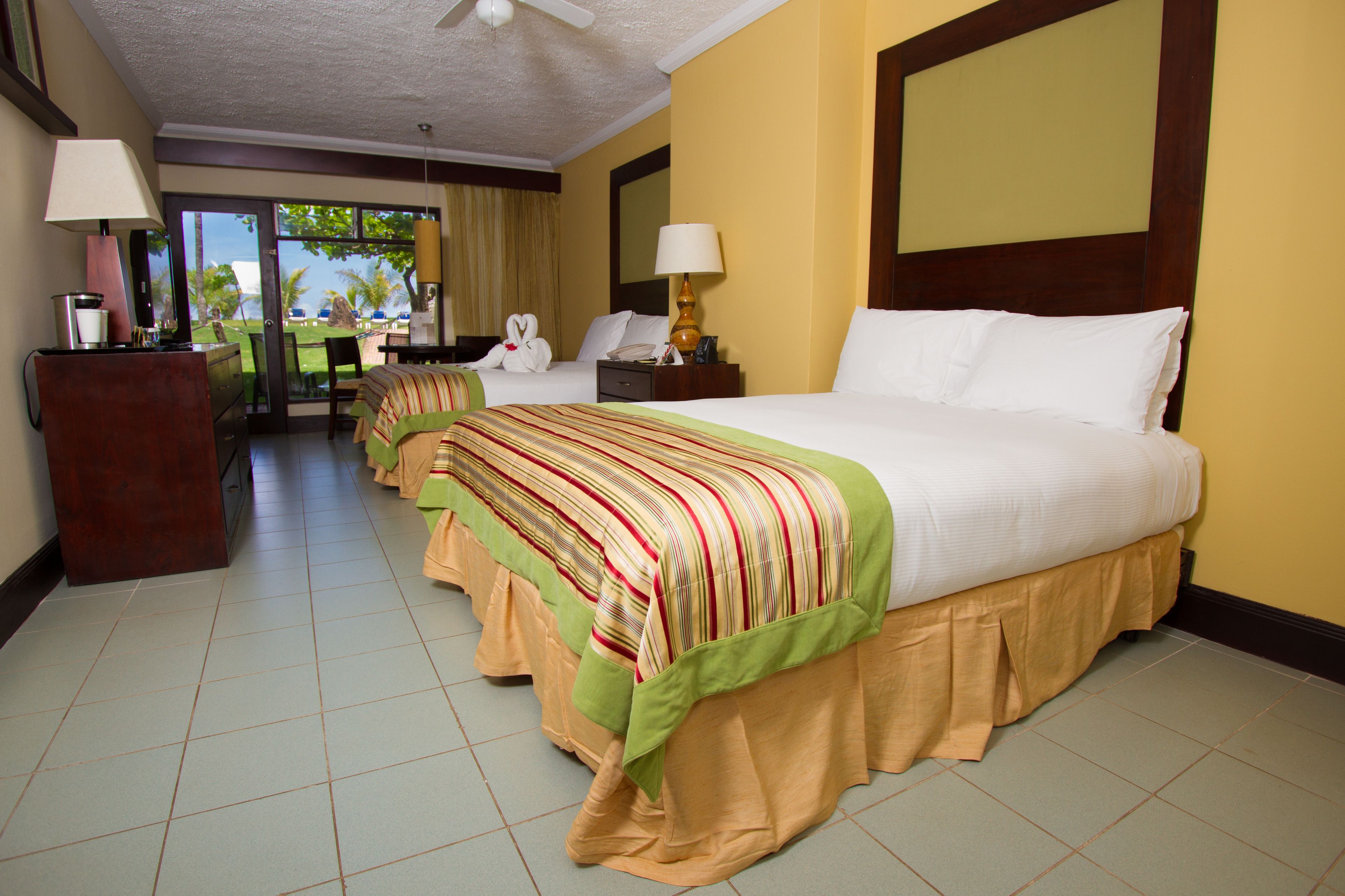 A hotel room with a large bed, a desk, and a chair. There is a window with a view of palm trees and a building.