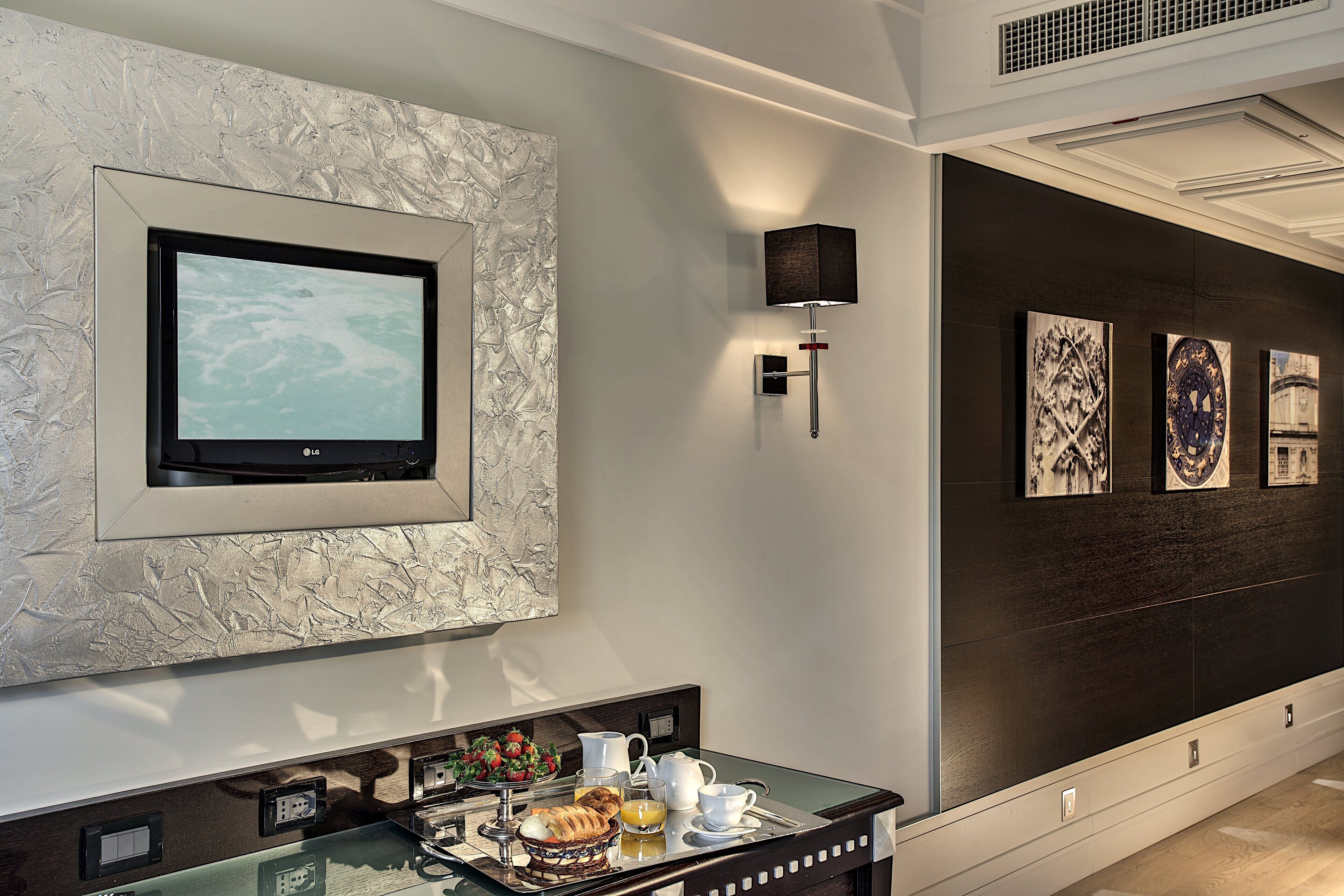A modern hotel room with a large flat-screen TV mounted on a textured wall, a tray table with breakfast items, and artwork on the dark wall.