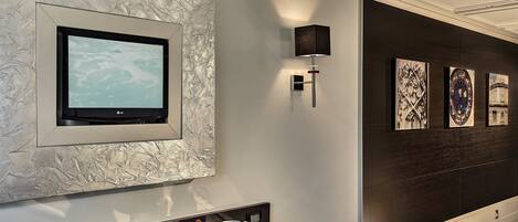 A modern hotel room with a large flat-screen TV mounted on a textured wall, a tray table with breakfast items, and artwork on the dark wall.