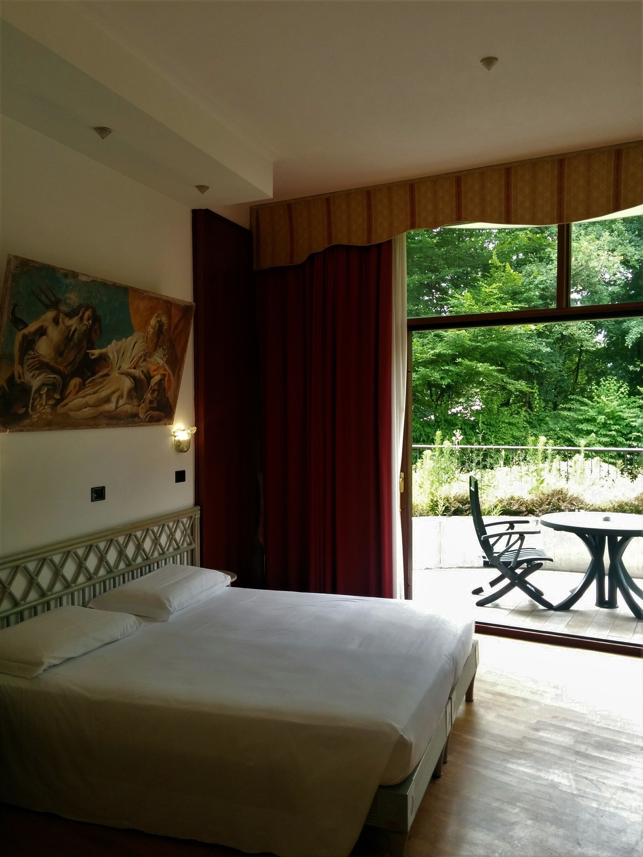 Habitación de hotel con cama, un cuadro en la pared, ventana con cortinas y balcón con mesa y sillas.