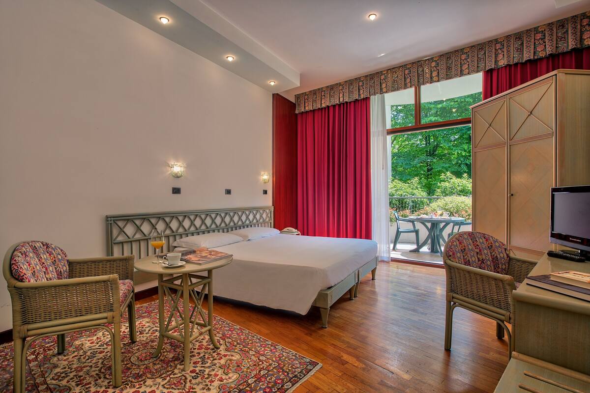 A hotel room with a bed, two wicker chairs, a small table, a television, and a balcony with a table and chairs.