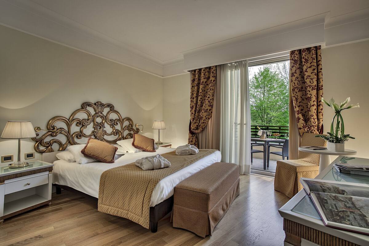 A hotel room with a large bed, two bedside tables, a bench, a balcony with a table, and a potted plant.