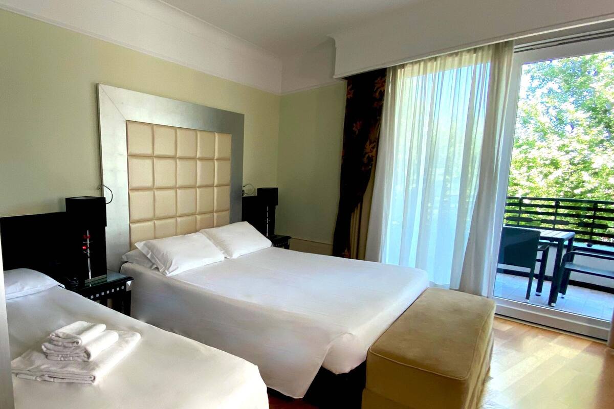 A hotel room with two beds, a wooden floor, a large window with curtains, and a balcony with outdoor furniture.
