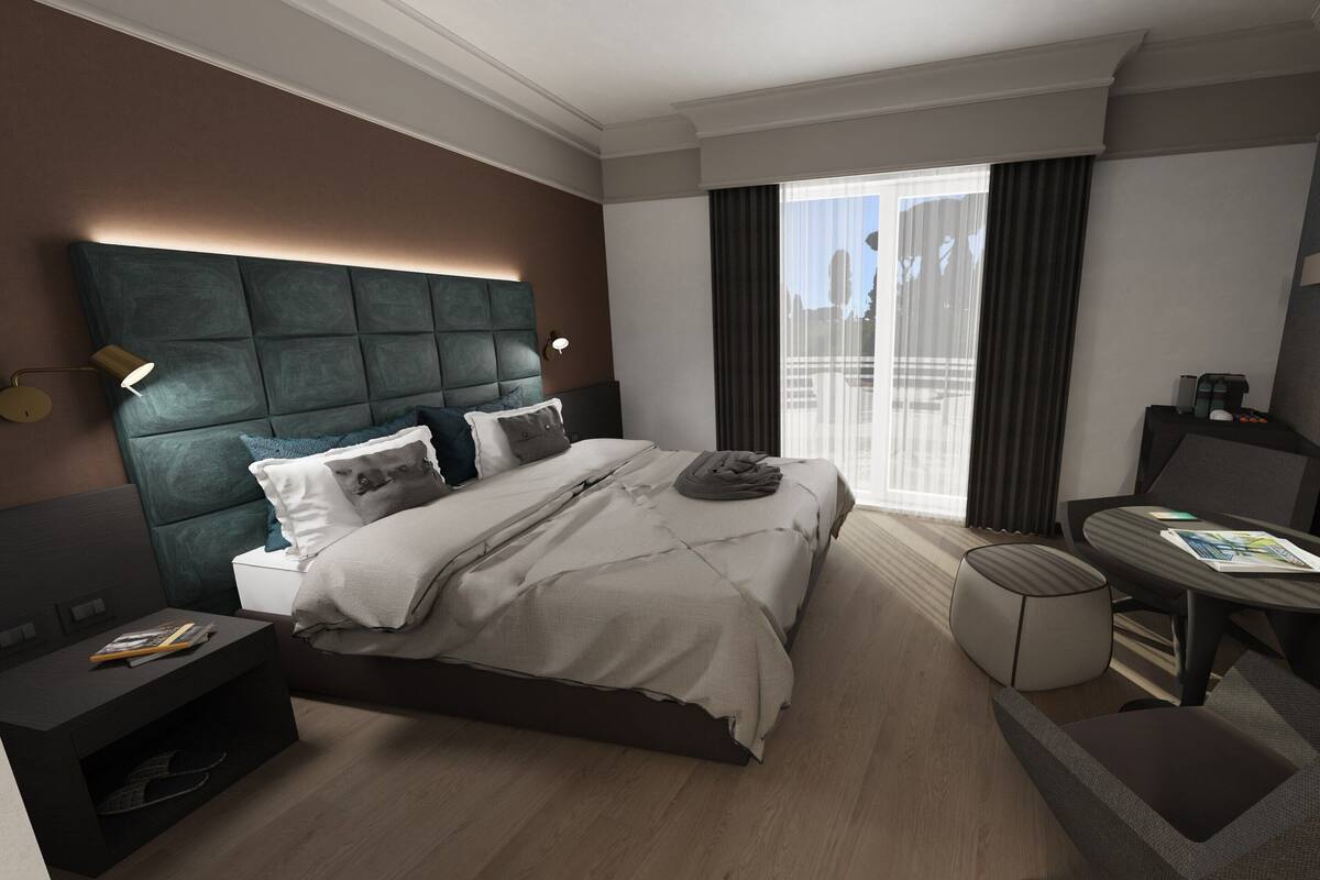 A modern hotel room with a large bed, a bedside table with a lamp, a small round table, and a large window with curtains.