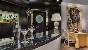 A bar area with a marble countertop, a cocktail shaker, and a martini glass. There is a round mirror on the wall and a painting of a horse. A table with a yellow tablecloth and a lamp is visible. - Park Hotel Junior (Quarto d'Altino)
