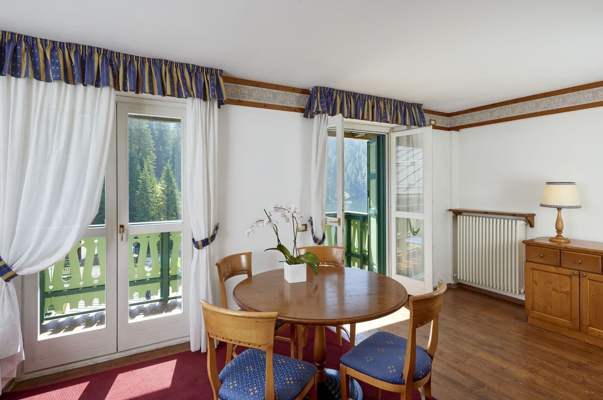 Een eethoek met een ronde houten tafel, blauwe fauteuils, een vaas met bloemen en uitzicht op een bos door de ramen.