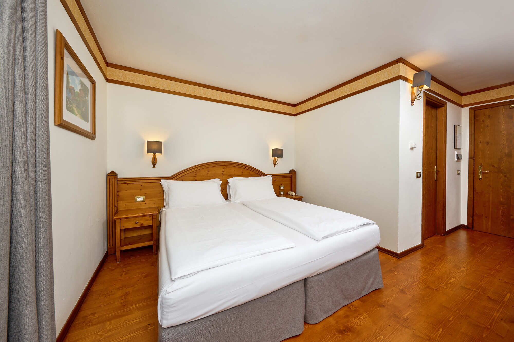 A hotel room with a double bed, wooden headboard, bedside tables, wall-mounted lamps, and a wooden door.