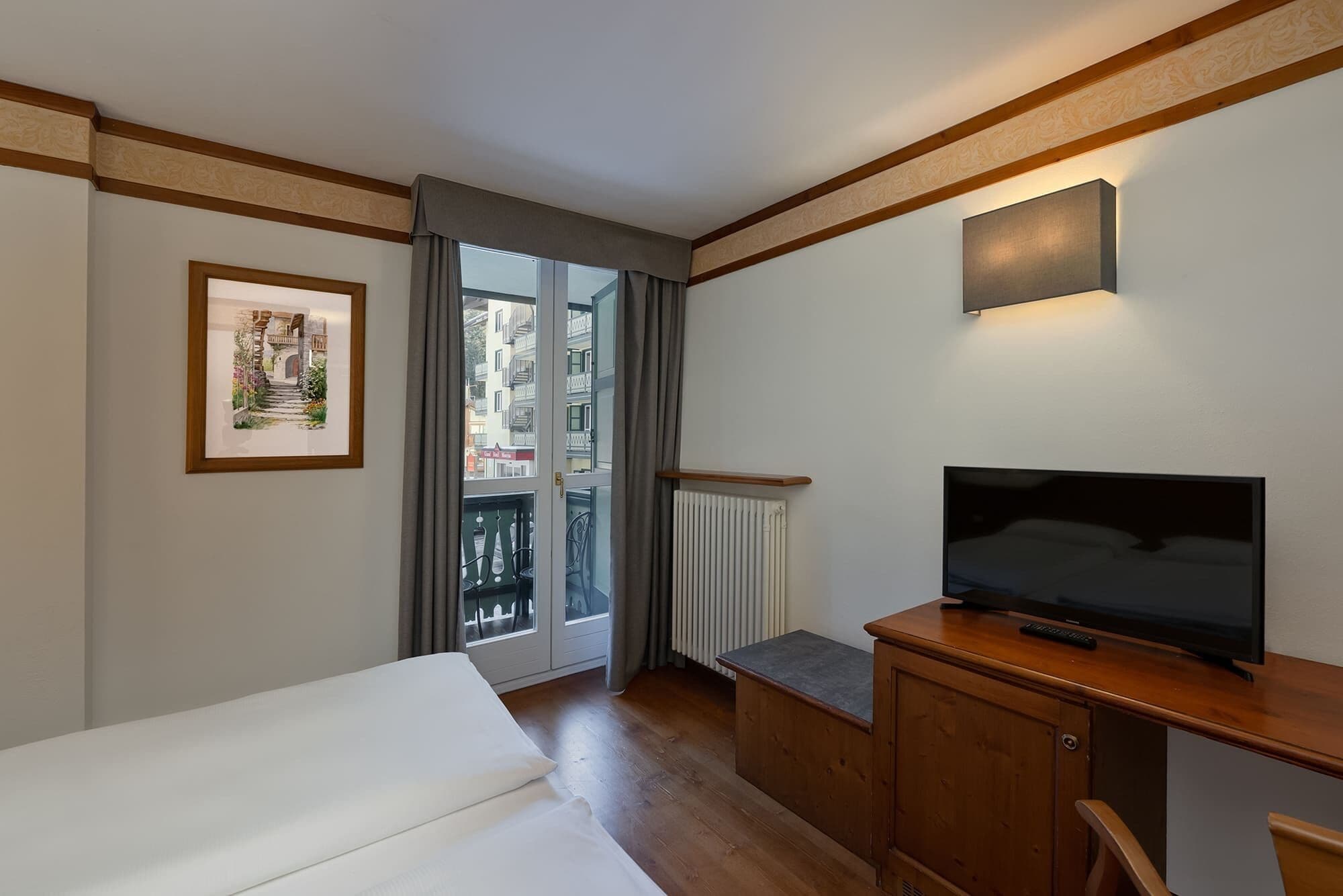 A hotel room with a bed, a TV on a wooden cabinet, a framed picture on the wall, and a window with a view of buildings.