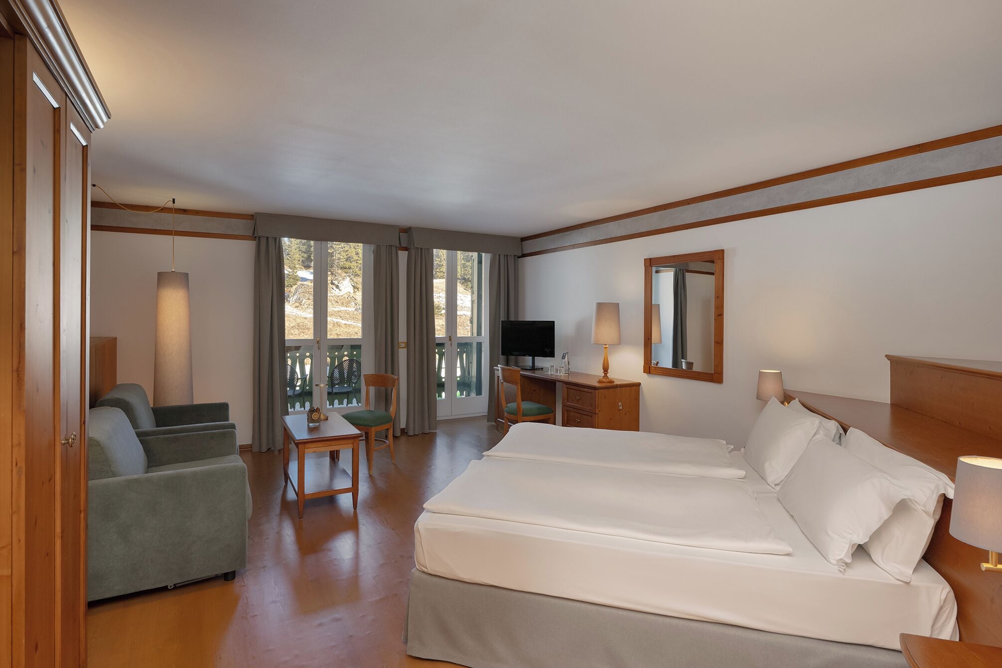 Habitación de hotel con cama, escritorio, silla, televisión y balcón con cortinas.