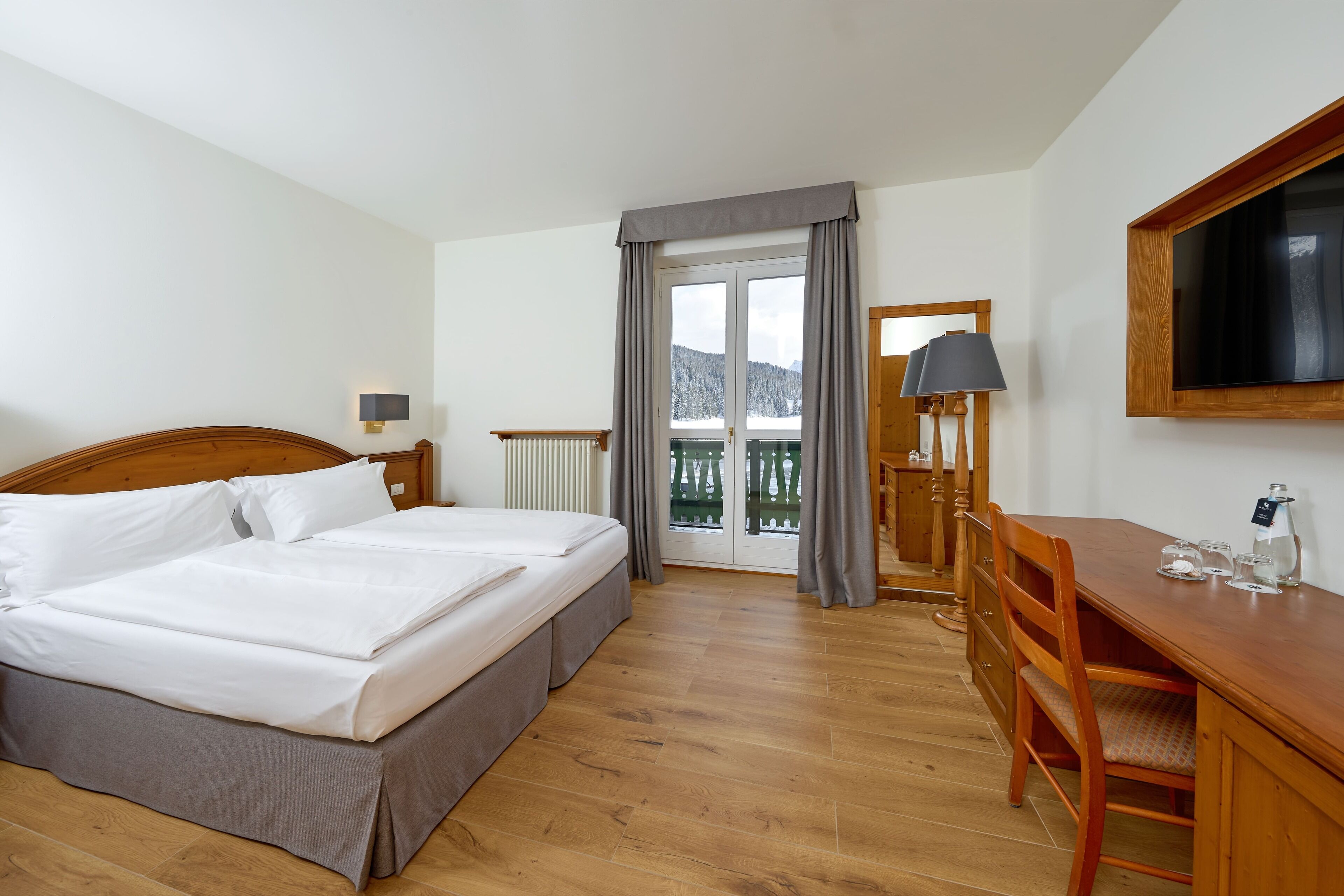 A hotel room with a double bed, a wooden desk, a chair, a TV, and a balcony with a view of the mountains.