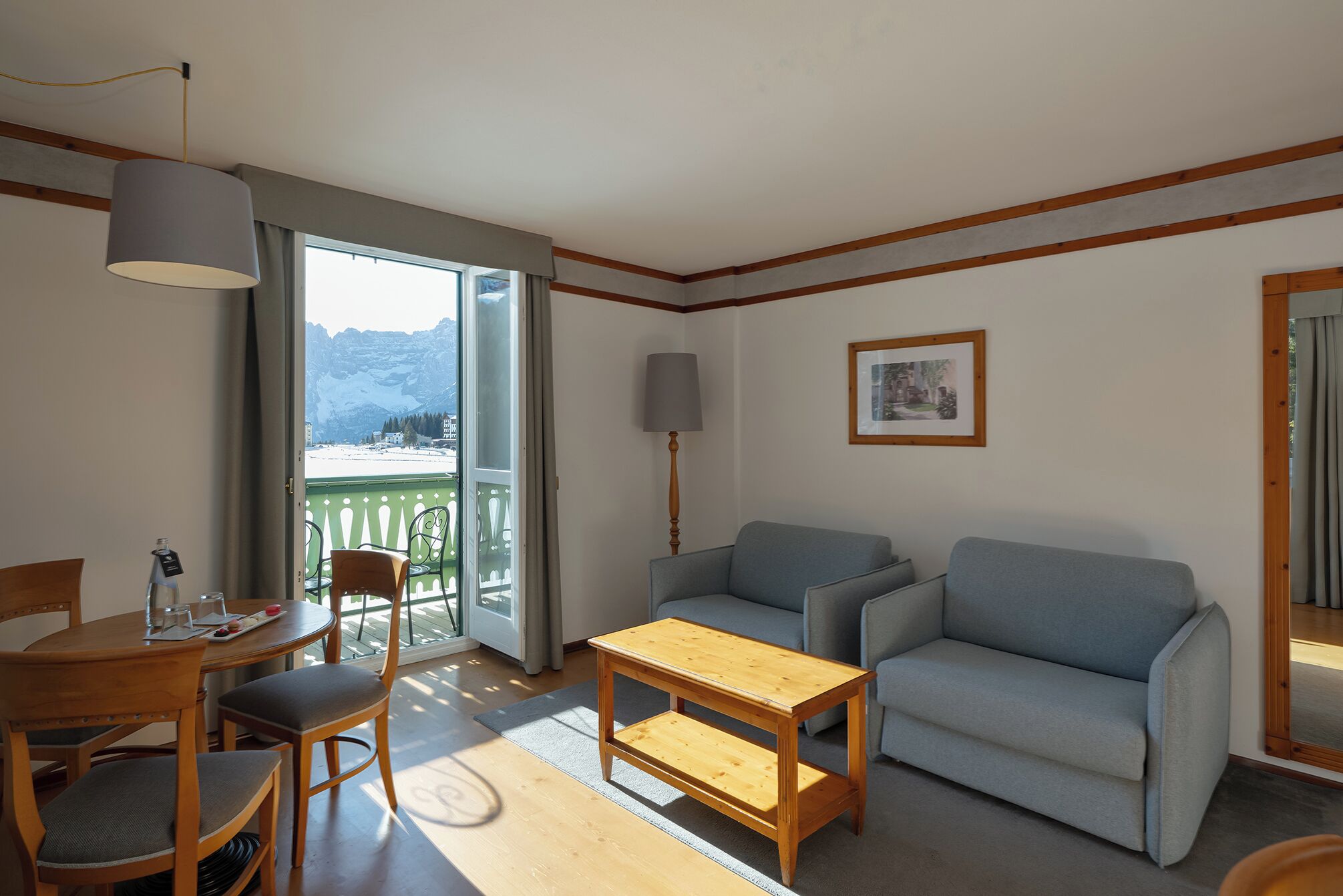 Une pièce chaleureuse avec une table à manger en bois, deux chaises, un canapé, une petite table basse en bois, une lampe sur pied et un balcon offrant une vue sur les montagnes.