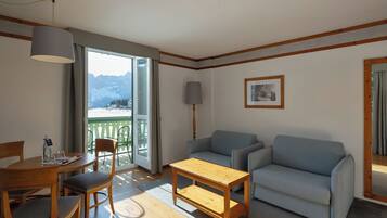 Une pièce chaleureuse avec une table à manger en bois, deux chaises, un canapé, une petite table basse en bois, une lampe sur pied et un balcon offrant une vue sur les montagnes.