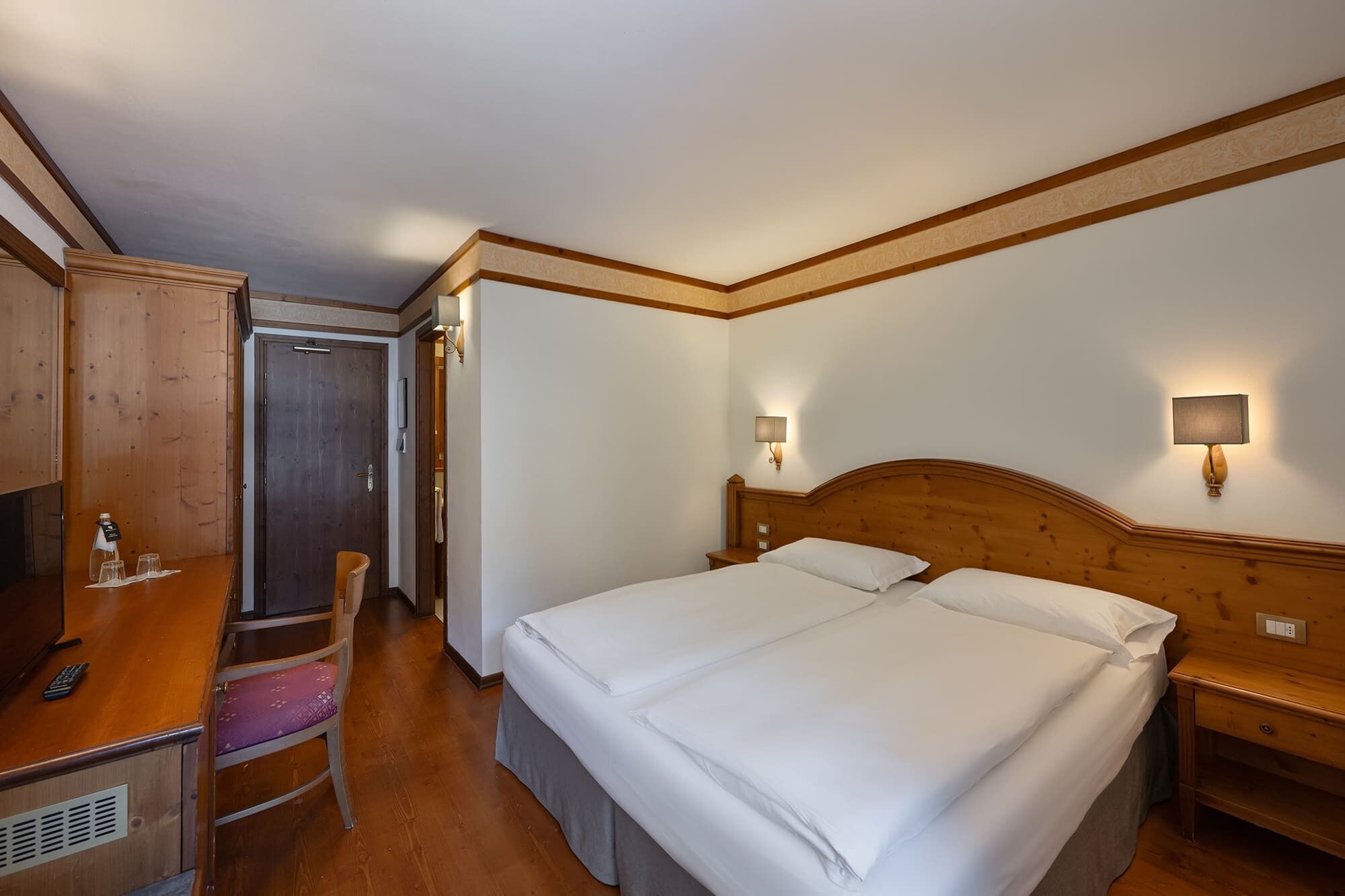 A hotel room with a wooden bed, a desk with a chair, a wardrobe, and a bathroom visible through an open door.