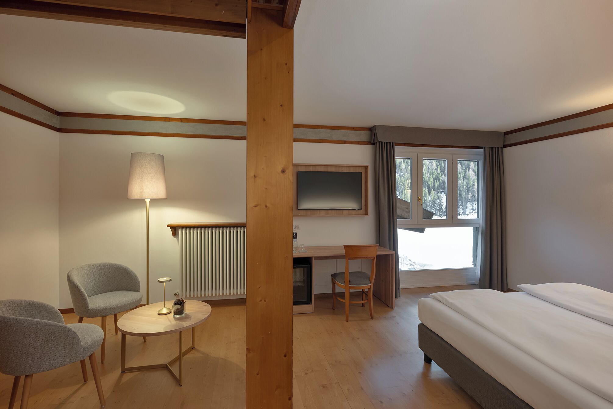 A hotel room with a bed, a desk, two chairs, a lamp, a radiator, and a window with curtains.