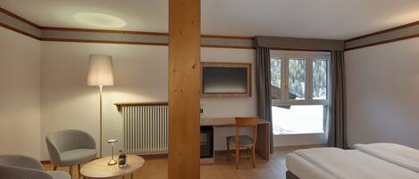 Habitación de hotel con cama, escritorio, dos sillas, lámpara, radiador y ventana con cortinas.