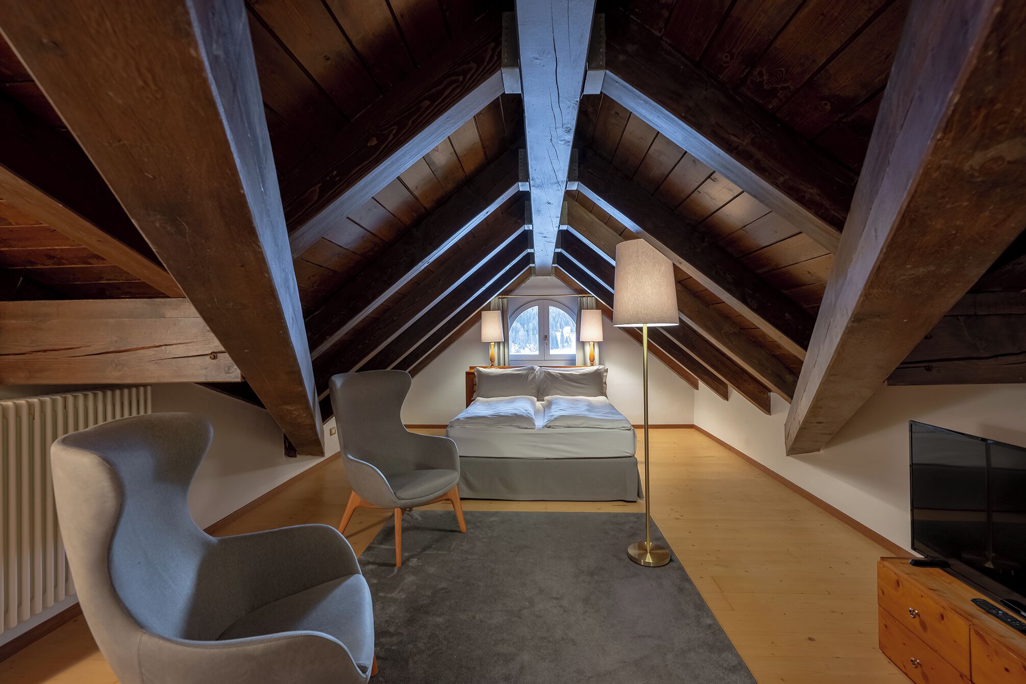 A bedroom with a sloped ceiling, a bed, a desk, a chair, a lamp, and a TV.