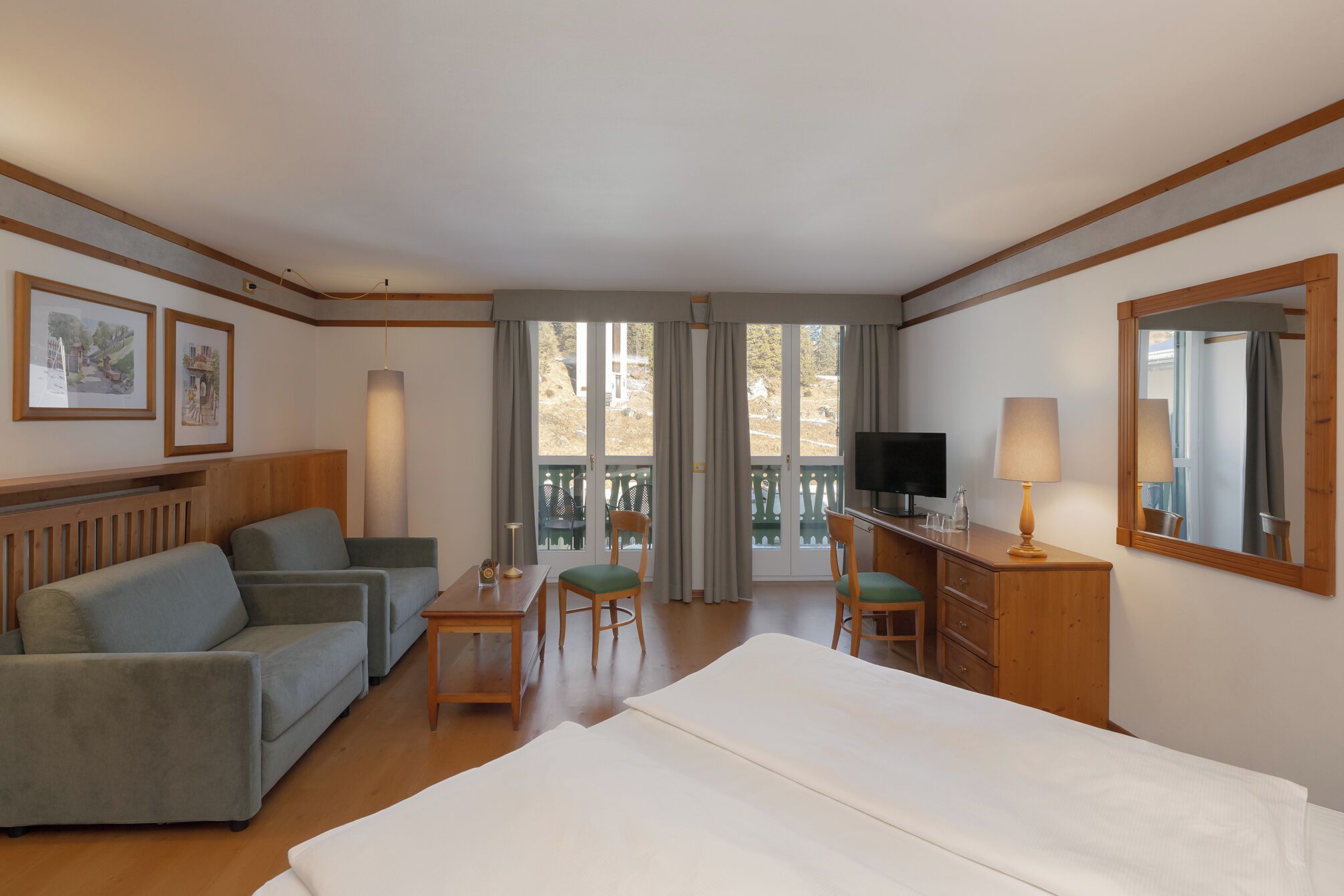 Hotelkamer met een bed, een bank, een bureau met lamp, een televisie en een balkon met stoelen.