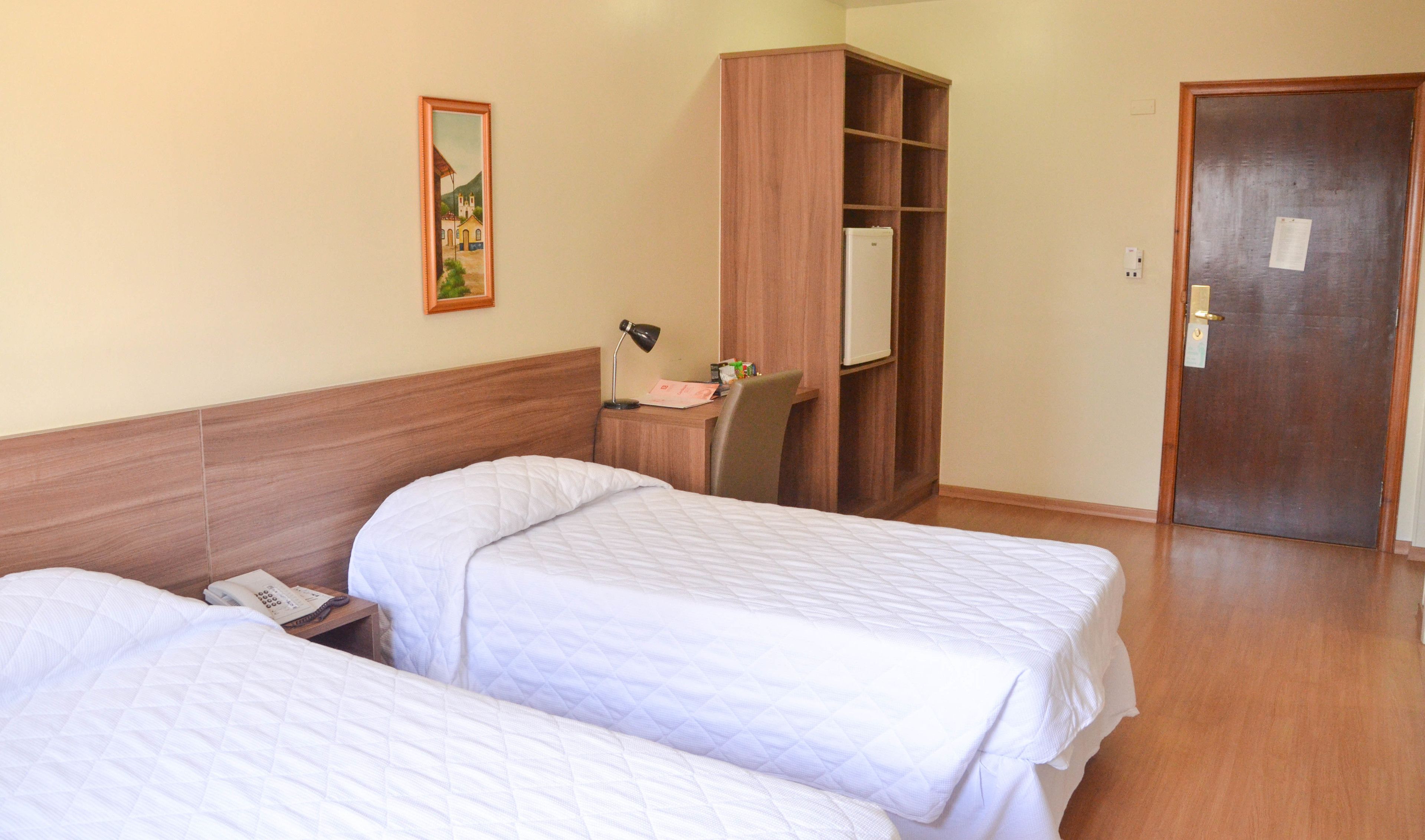 A hotel room with two beds, a wooden wardrobe, a desk with a chair, a desk lamp, and a painting on the wall.